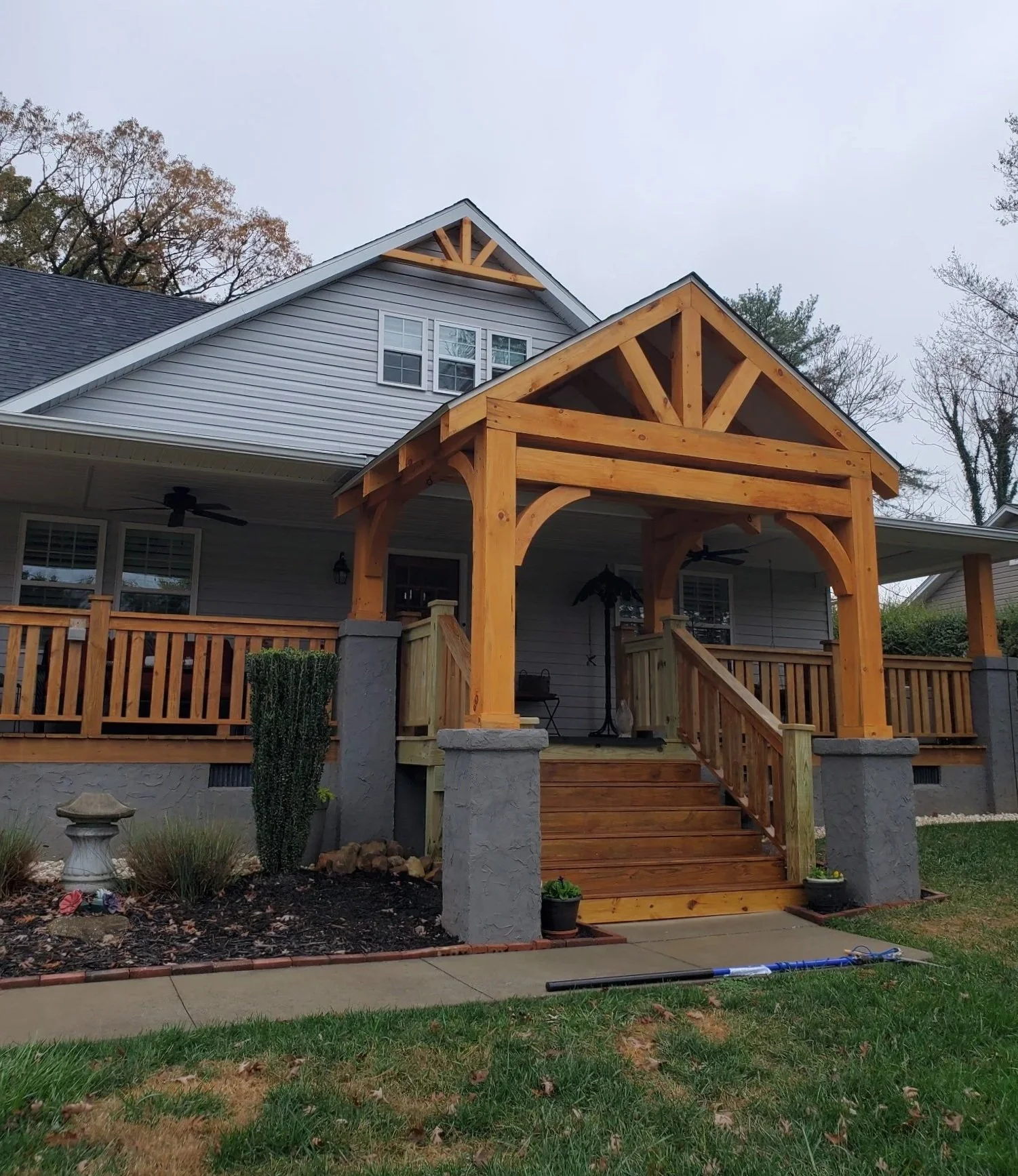 Front porch of a house with a newly built wooden pergola supported by stone pillars, stairs leading up to the porch, and a landscaped yard in the foreground.