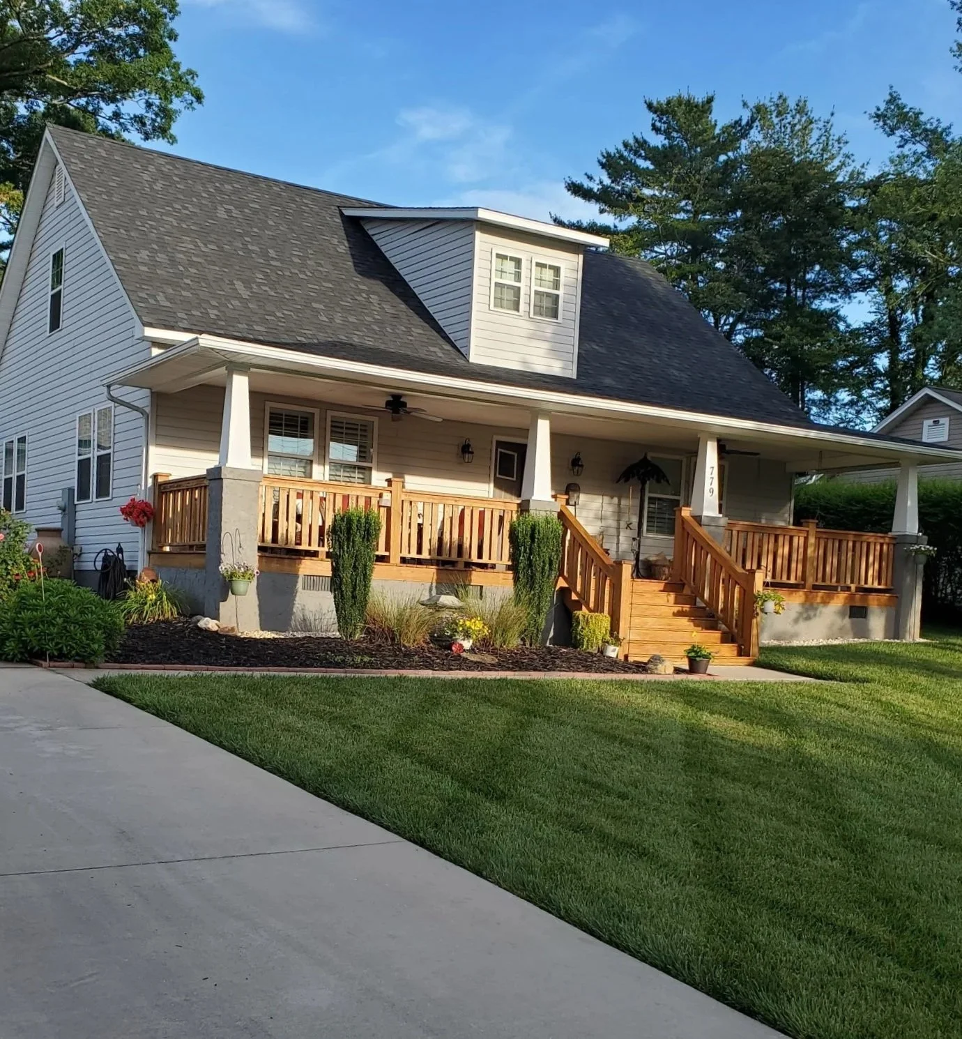 Front view of a two-story house with a gray roof, white siding, a covered porch with wooden railings and stairs, landscaped yard with green grass, small plants, and trees, under a blue sky.