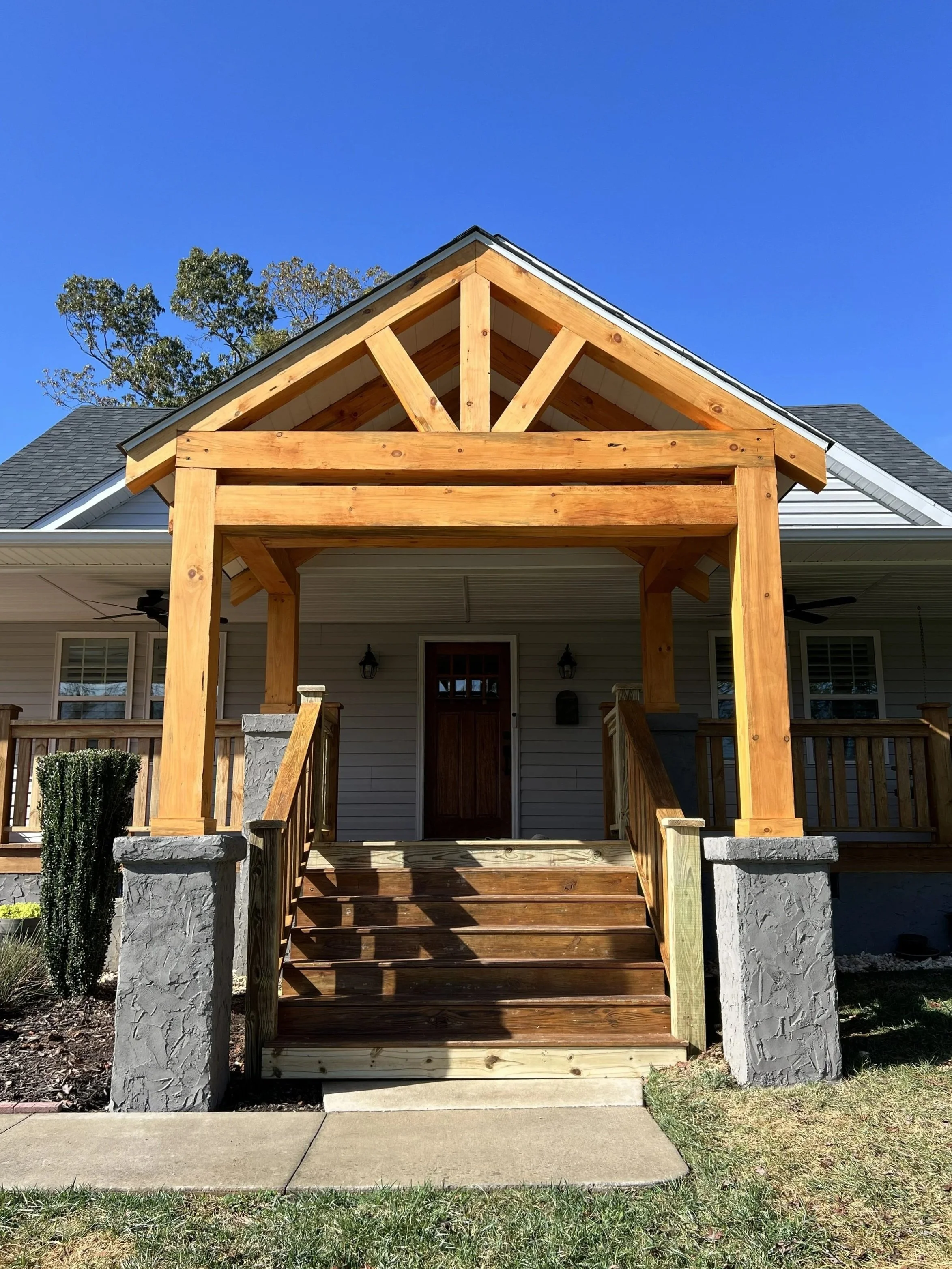 Front porch of a house under construction with wooden beams and steps, painted stone pillars, and a wooden door, on a sunny day with clear blue sky.