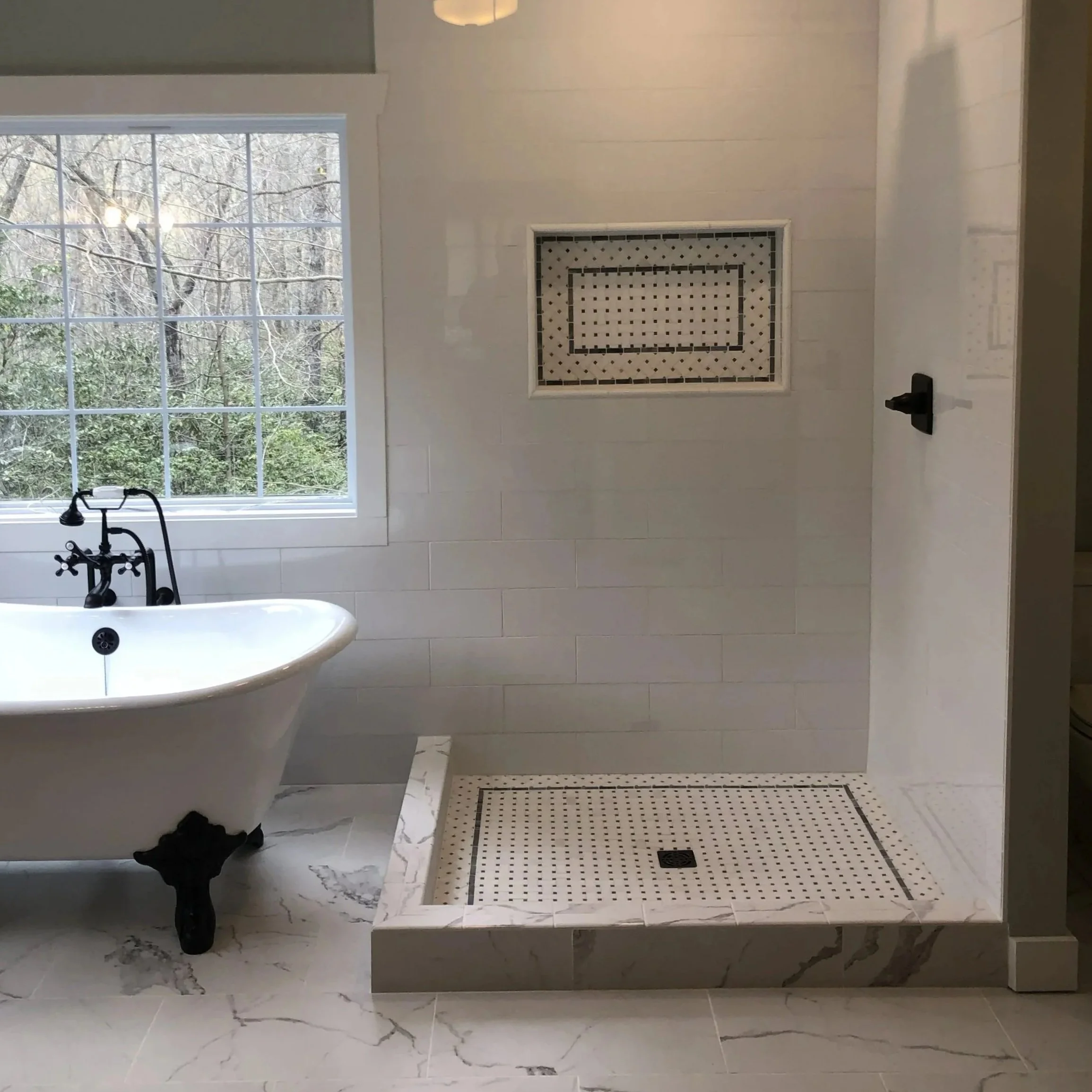 Bathroom featuring a clawfoot bathtub next to a large window, with a walk-in shower on the right, tiled in white with a black and white patterned niche and shower floor.
