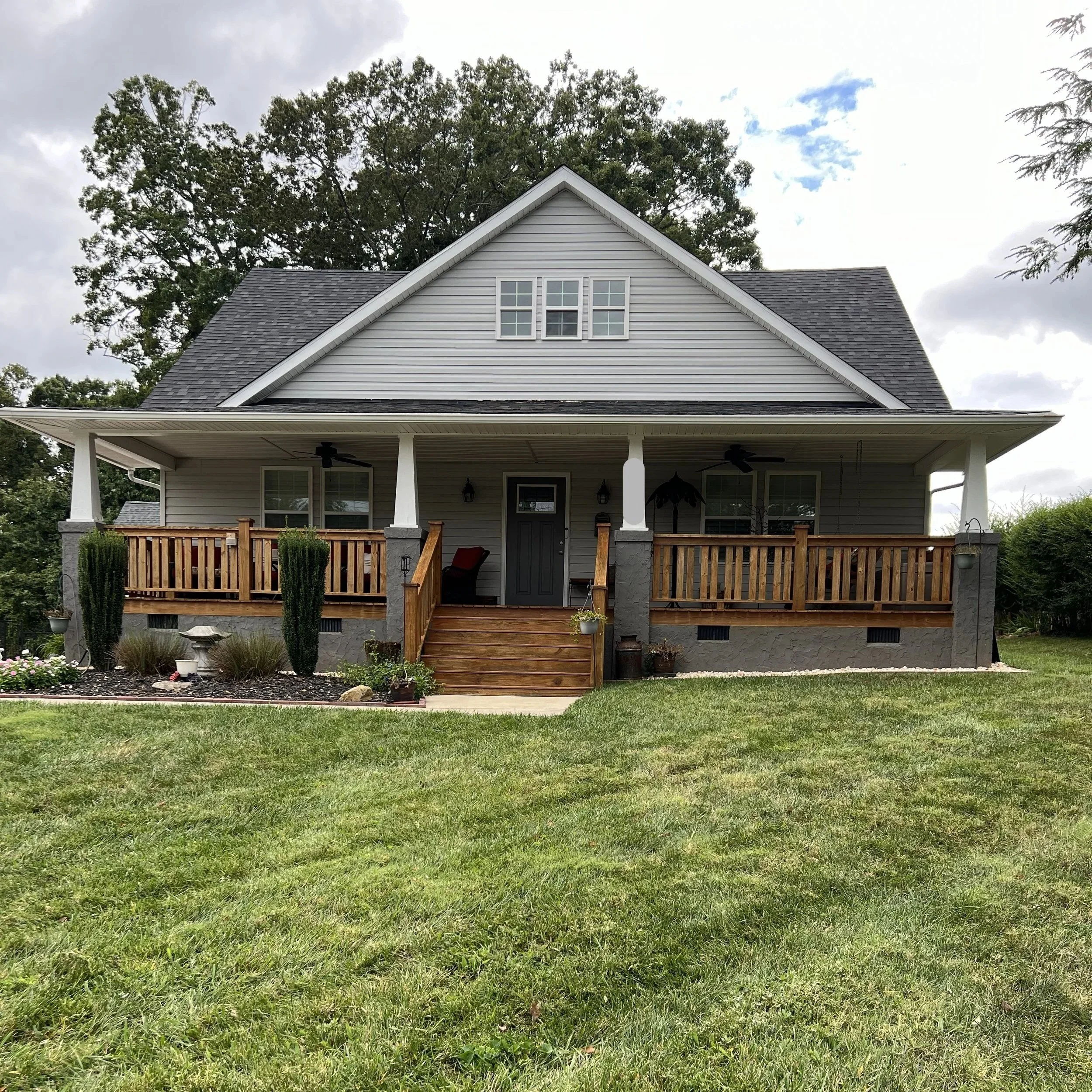 A two-story house with gray siding, a dark gray roof, and a covered porch with wooden railings and stairs, surrounded by a green lawn and trees.
