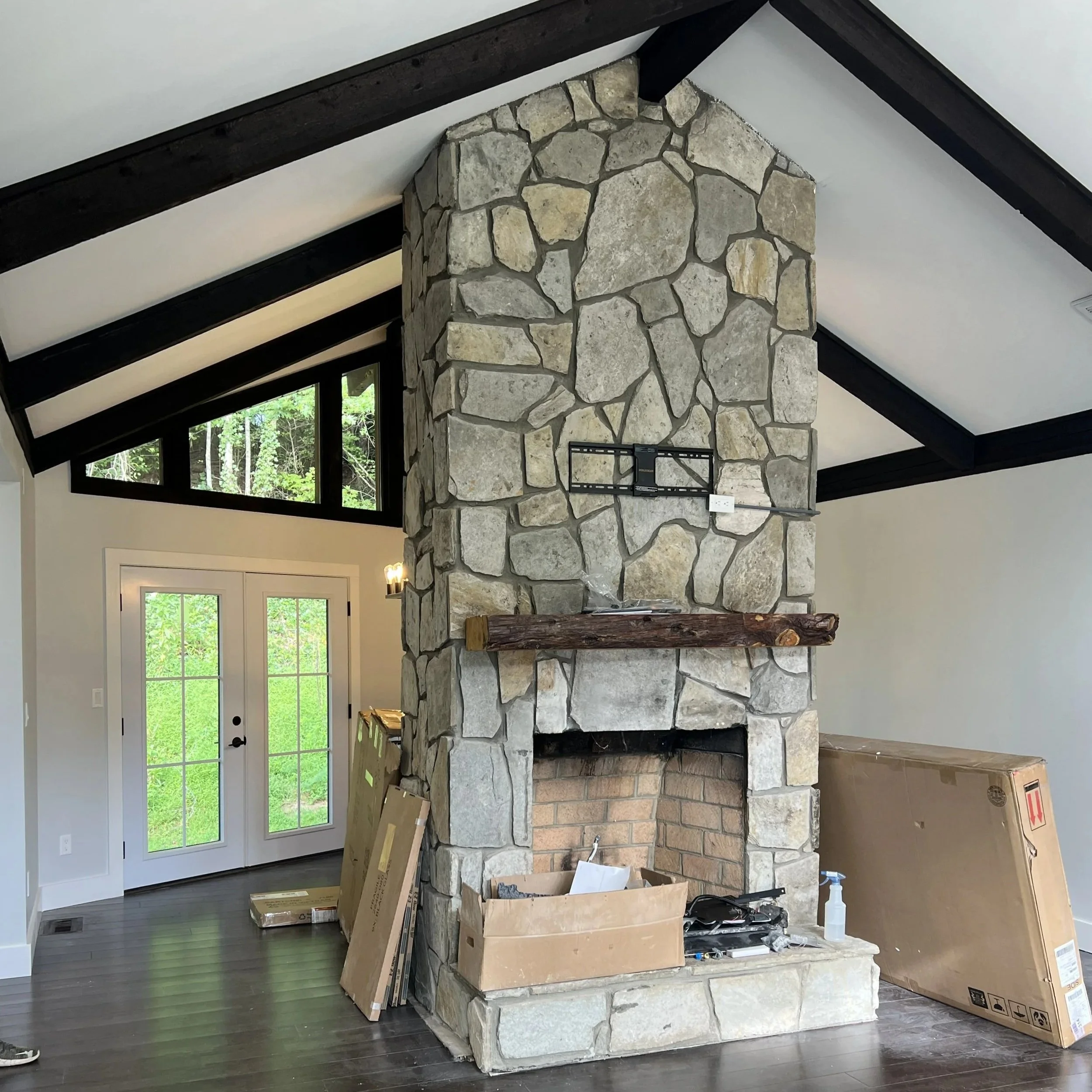 Interior of a house with a large stone fireplace, high vaulted ceiling with black beams, and glass double doors leading outside, with construction materials around.