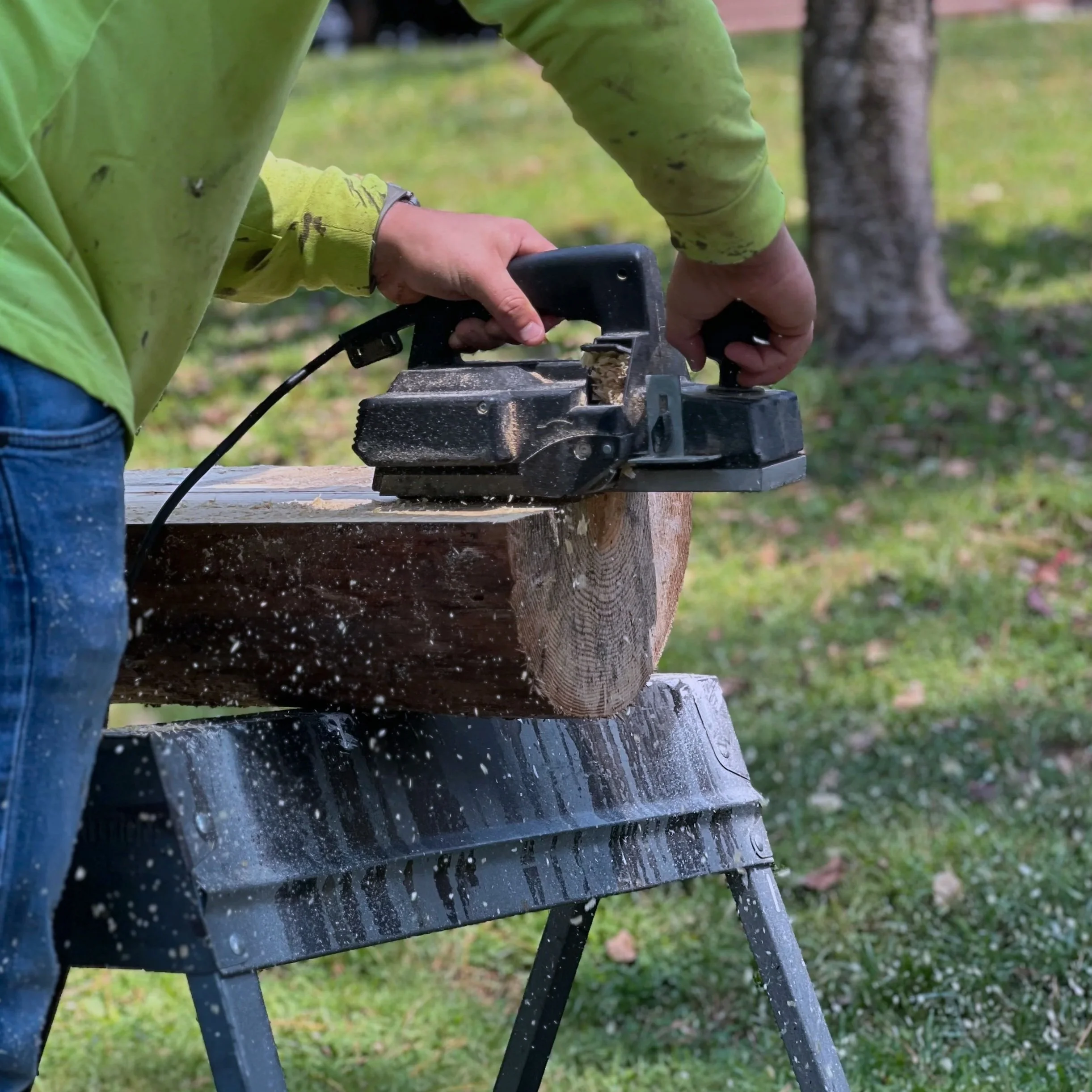 A person using a jigsaw to cut a piece of wood outdoors on a workbench, with sawdust flying around.