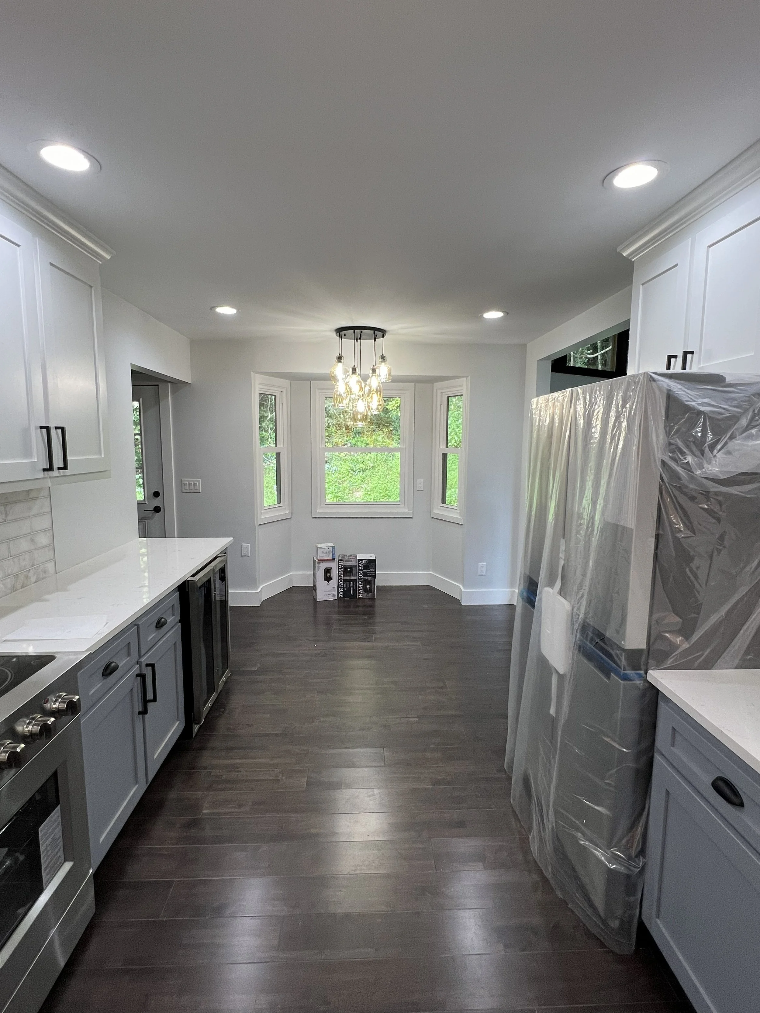 Empty kitchen with white cabinets, dark hardwood floors, and a window bay with three windows showing greenery outside. A modern chandelier hangs over the space, and some boxes are on the floor by the window.