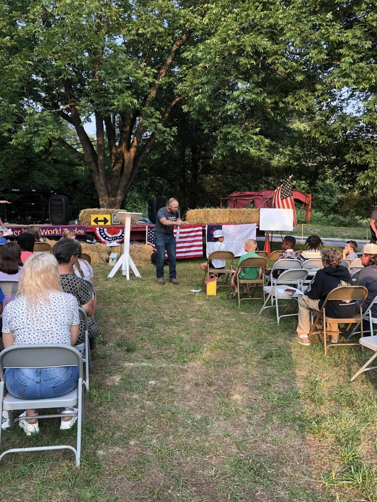 An outdoor event with a seated audience facing a man speaking into a microphone on a small stage decorated with American flags and patriotic banners, under a large tree with hay bales and a red barn in the background.