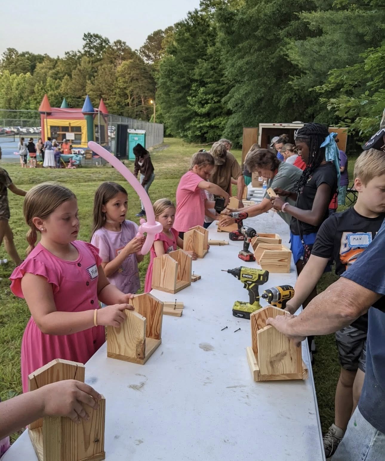 Children and adults building wooden birdhouses at an outdoor woodworking event and playground in the background.