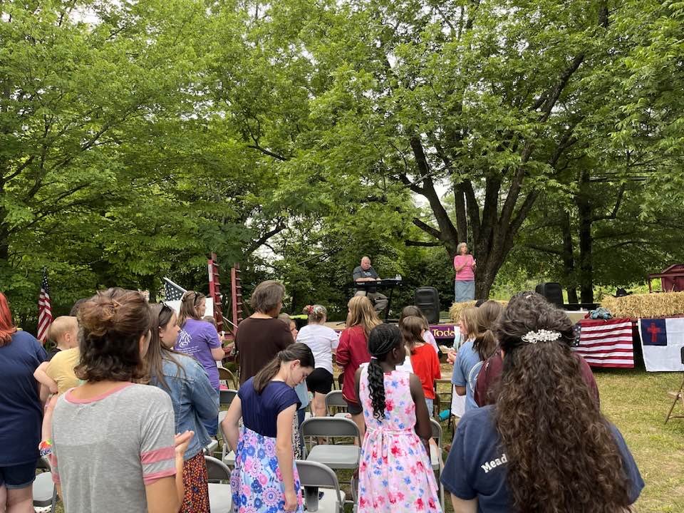 People gathered outdoors for a patriotic event with American flags and banners, watching a performance on a small stage under trees.