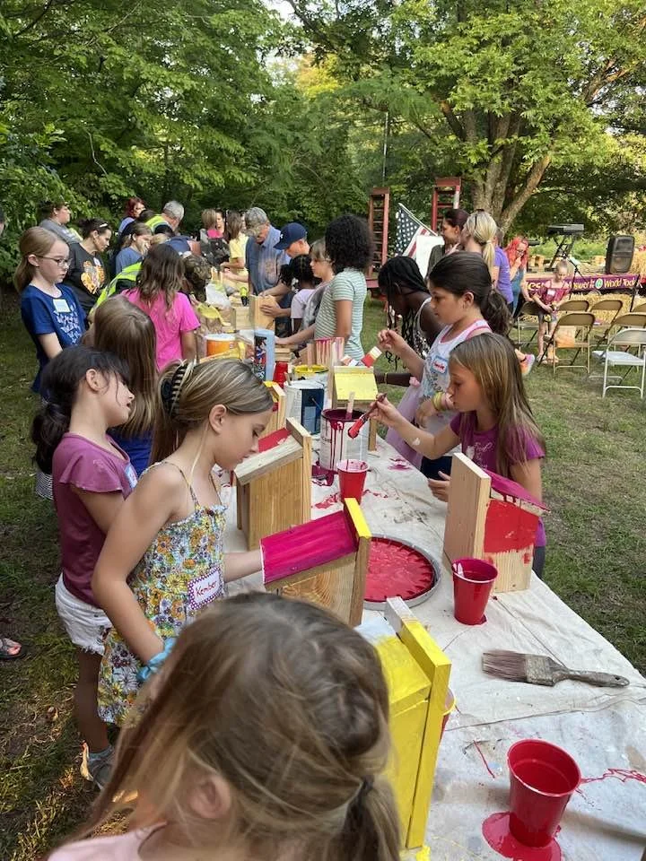 Children and adults participating in a craft activity outdoors, painting on wooden boxes, with trees and more people in the background.