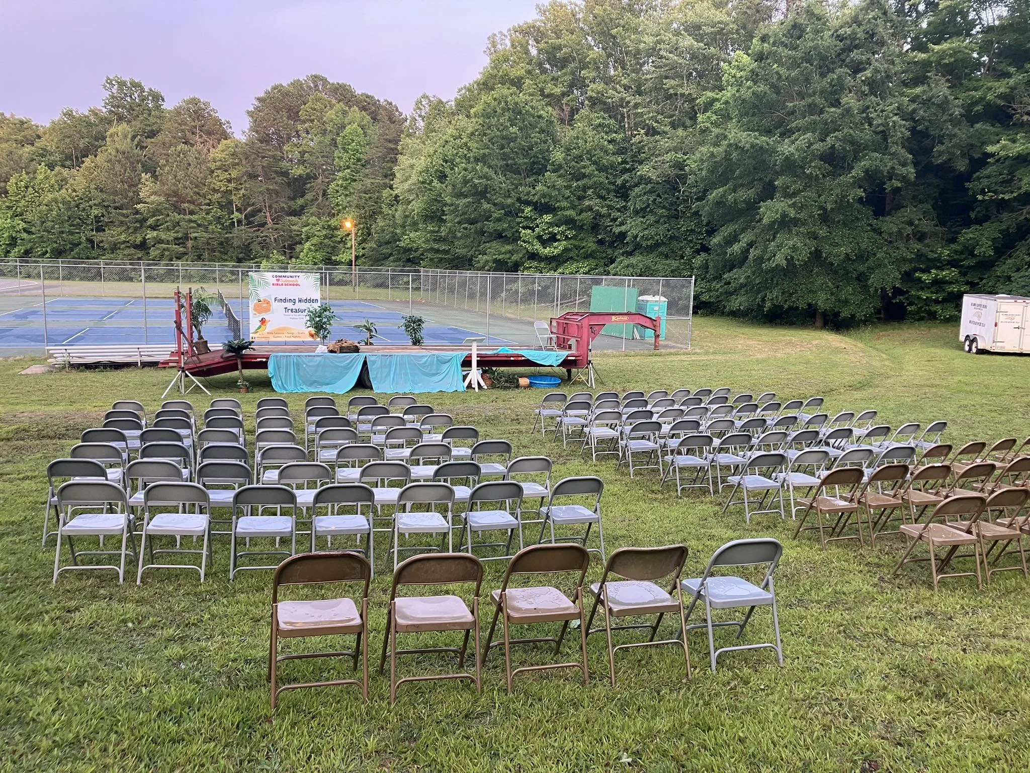 Outdoor event setup with rows of chairs facing a small stage with a banner, near a fenced tennis court, with trees in the background.
