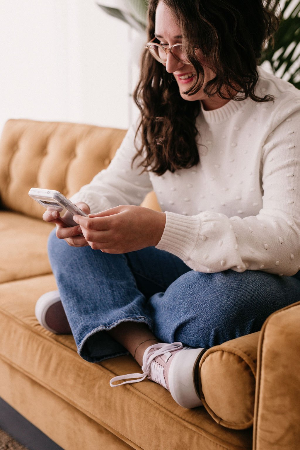 Femme assise sur un canapé beige, regardant son téléphone portable et souriant.