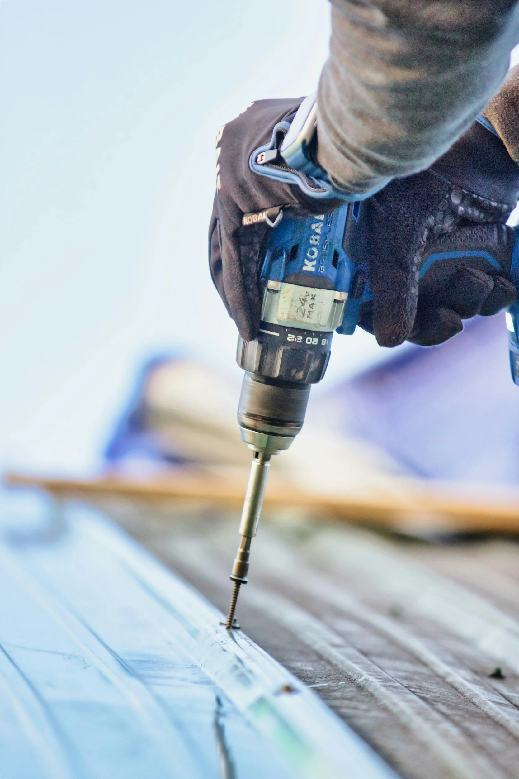 Close-up of a person wearing gloves using a power drill to fasten a screw into a wooden surface.