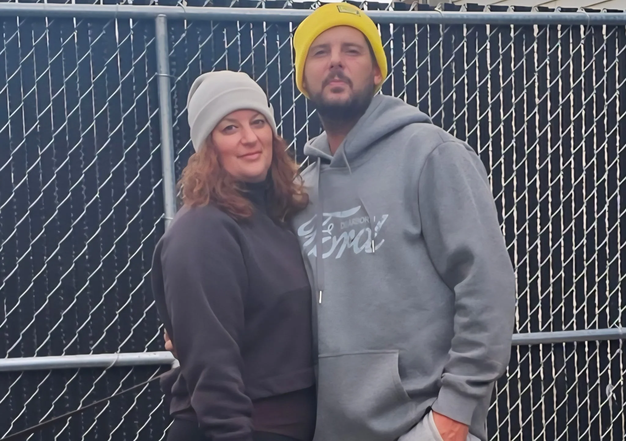 A man and woman standing close together outdoors, both wearing beanies and casual hoodies, in front of a chain-link fence.