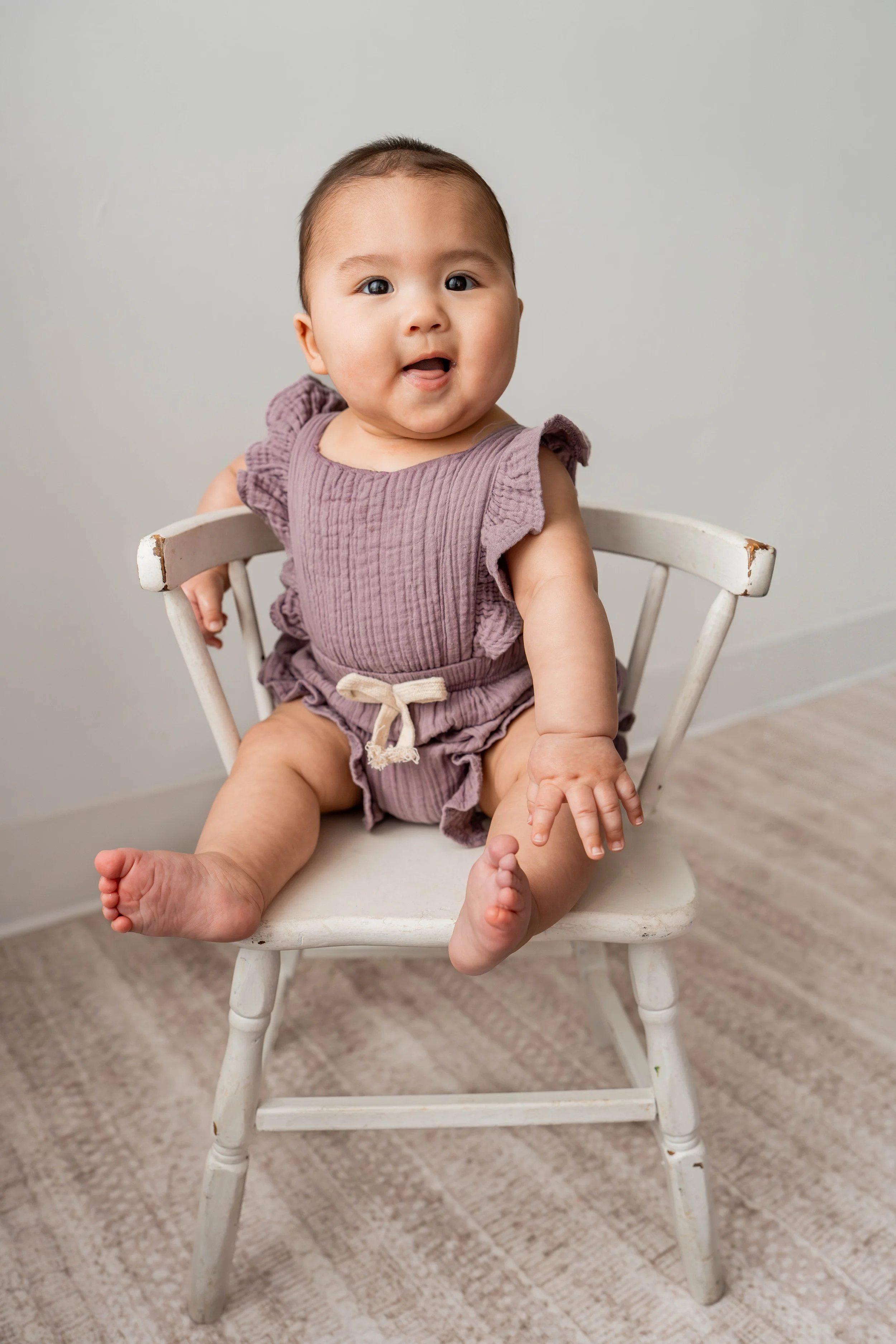 A baby sitting on a white wooden chair, wearing a purple ruffled romper, looking at the camera with a happy expression.