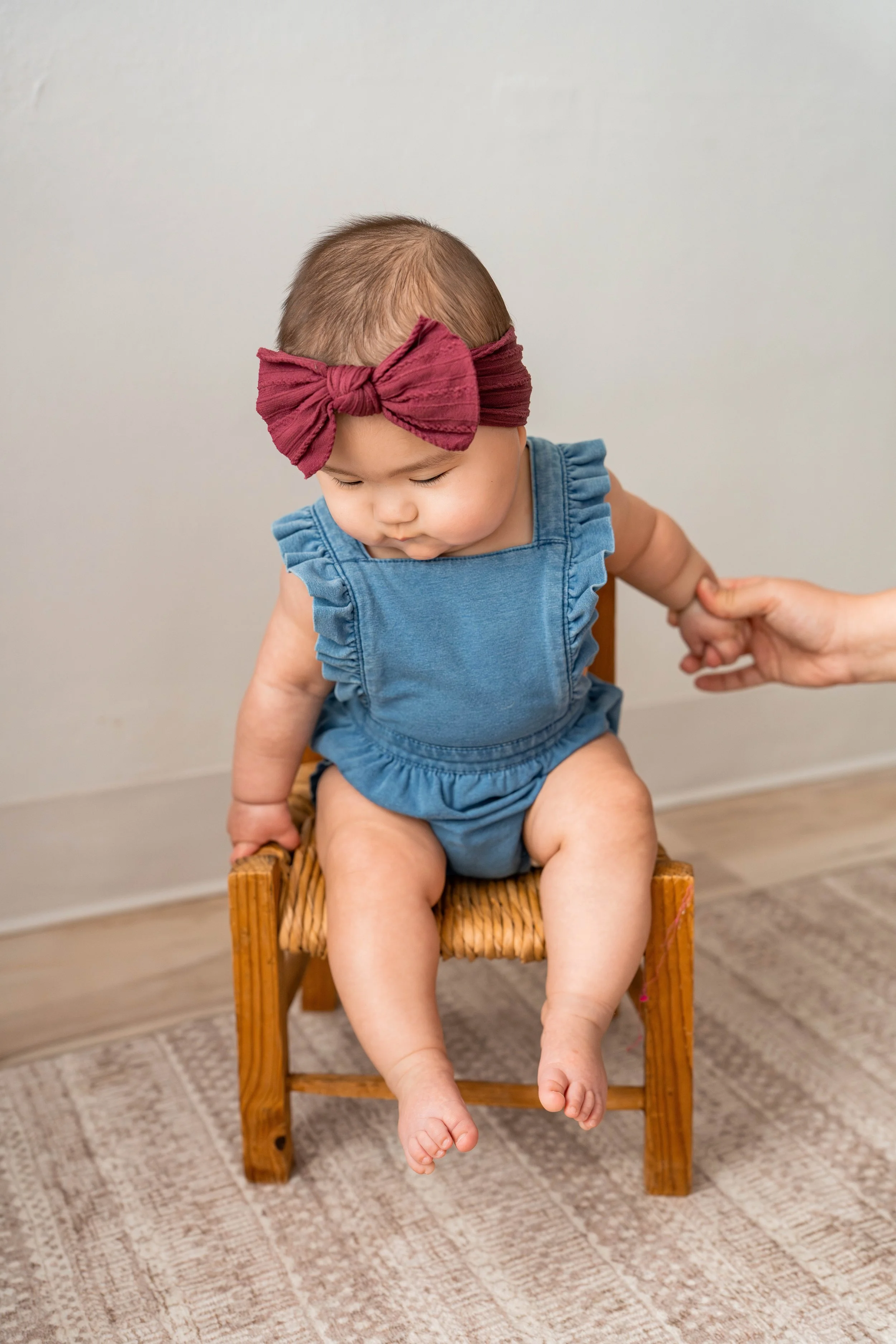 Baby girl in jean overalls and a burgandy bow sitting in a chair 