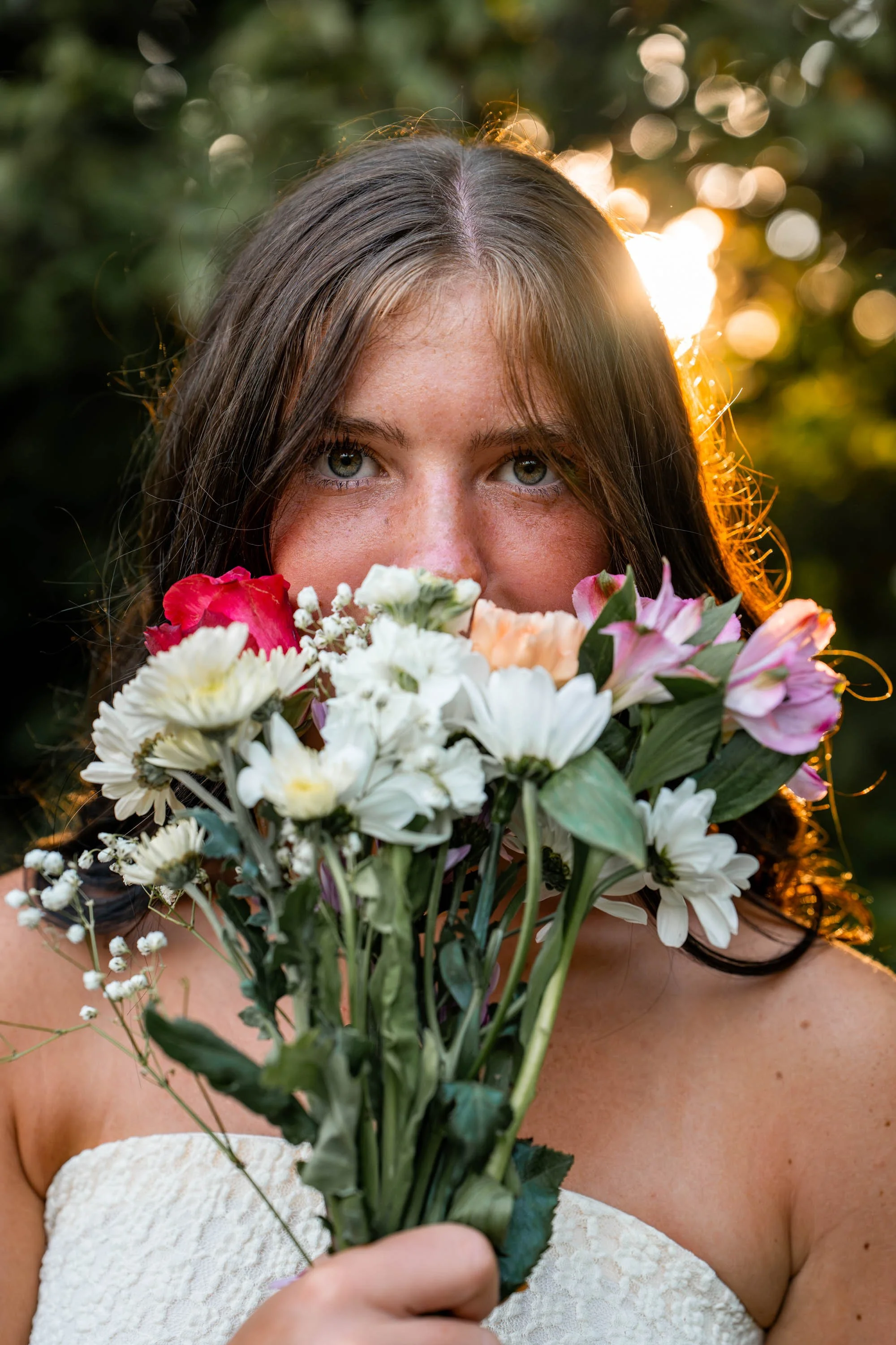 a girl stares into the camera holding flowers in front of her face while the sun shines in through the trees