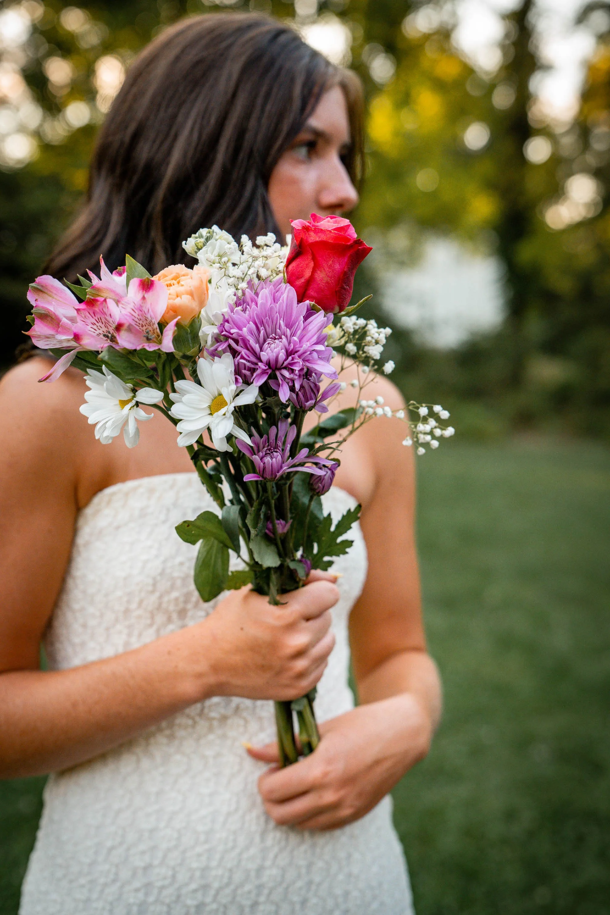 a senior girl looks off into the distance while holding a bouquet of flowers and wearing a white dress