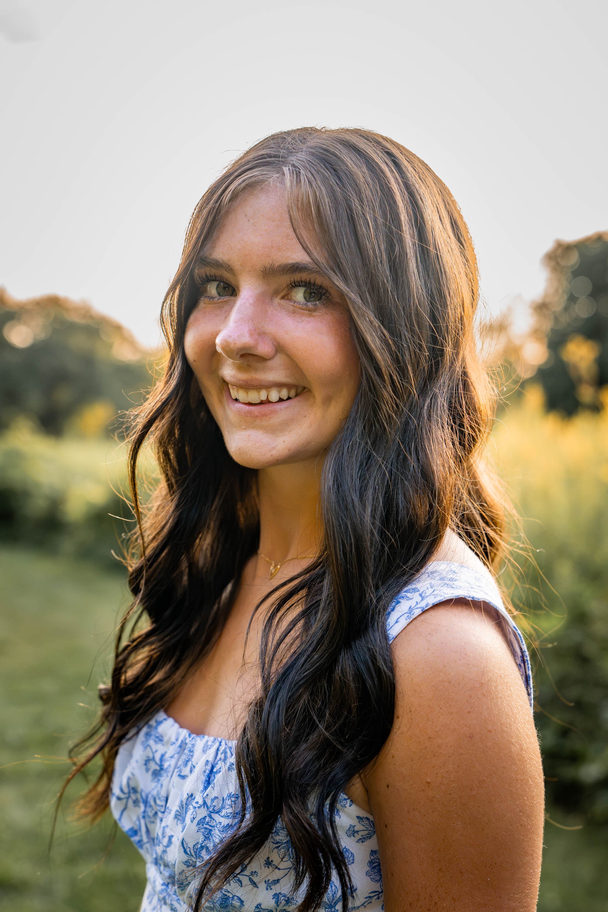 a girl in a sleeveless dress smiles at the camera with her wavy brown hair falling over her shoulders