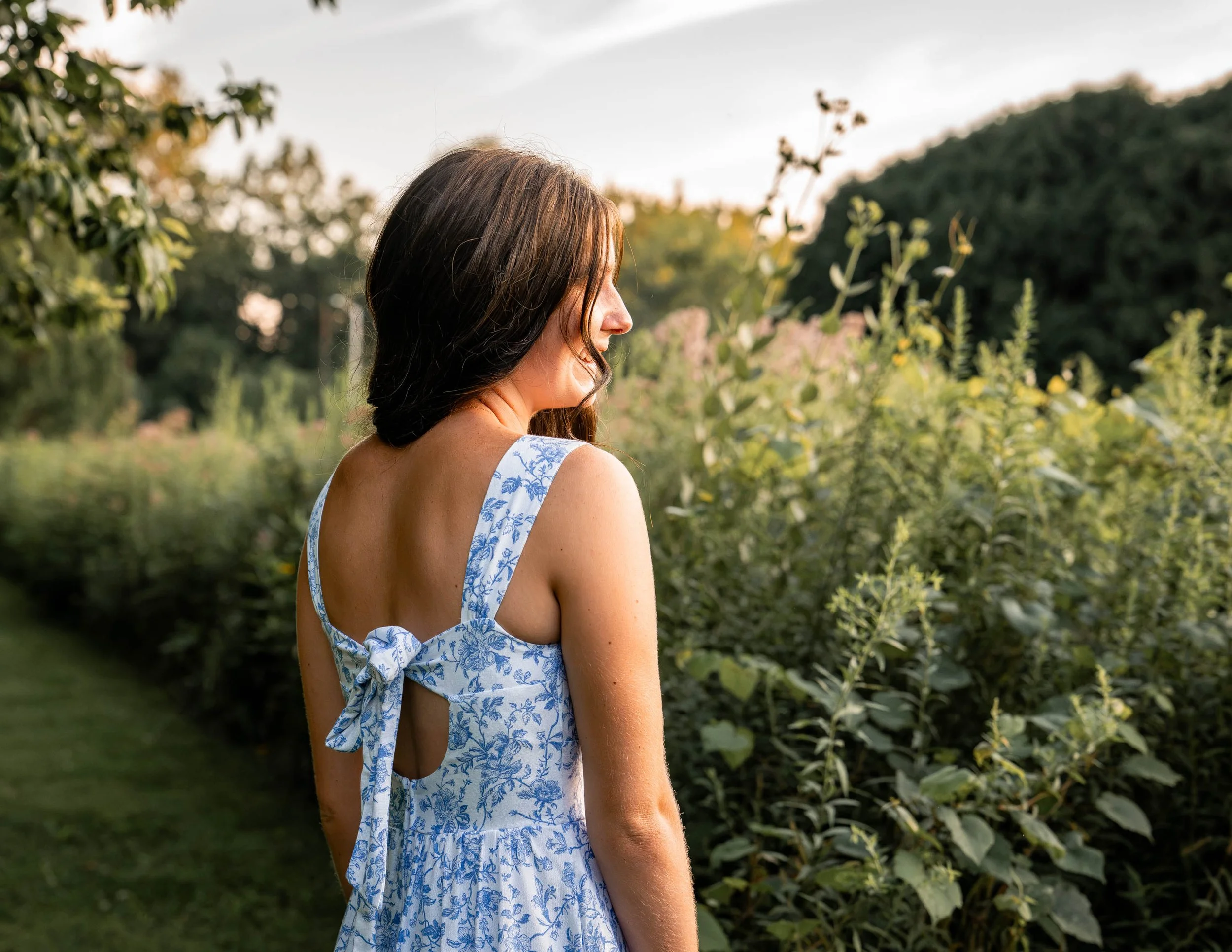 a girl looks out at the wildflowers while wearing a blue and white dress showing the bow on the back