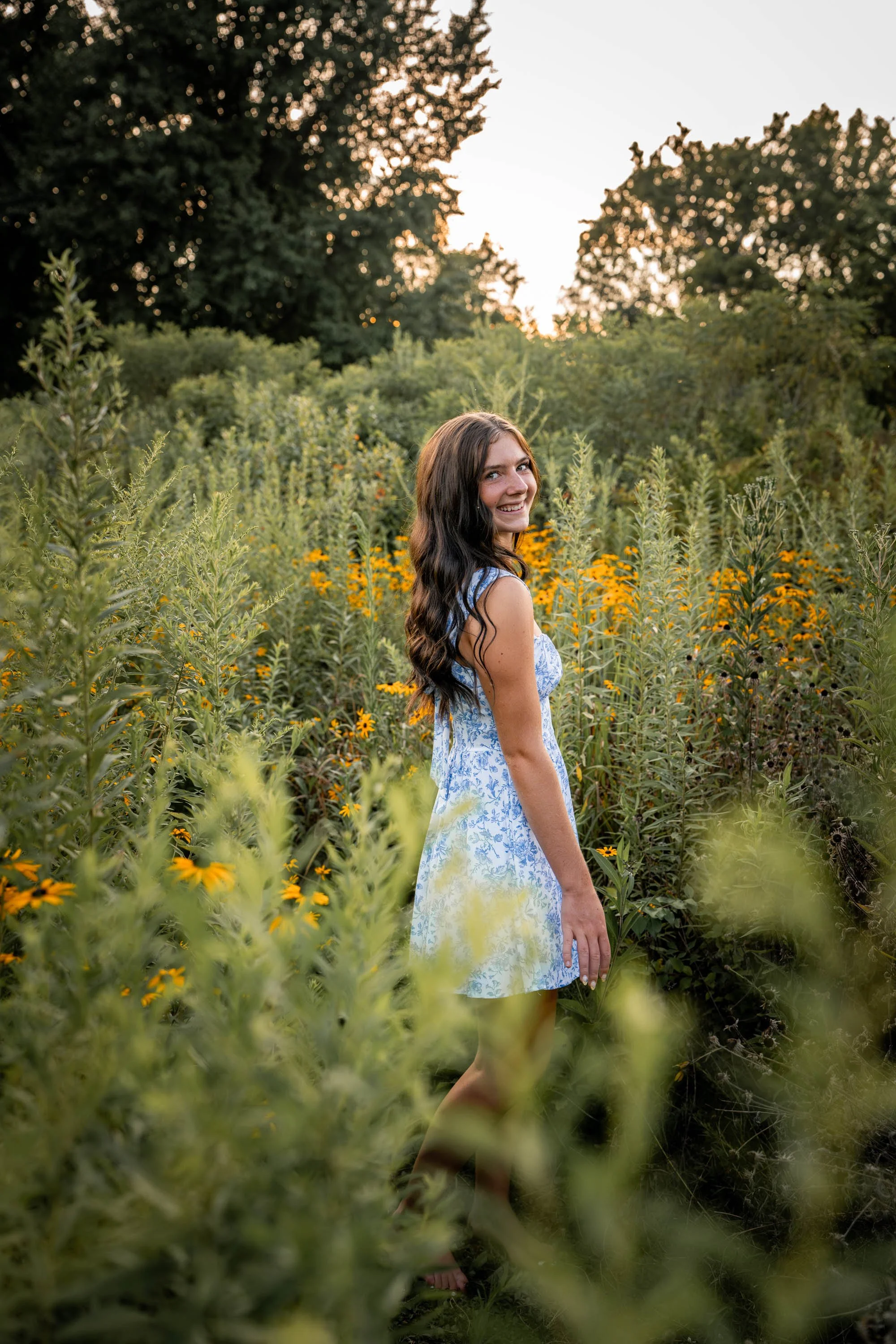 senior girl looks back as she walks through a field of wildflowers