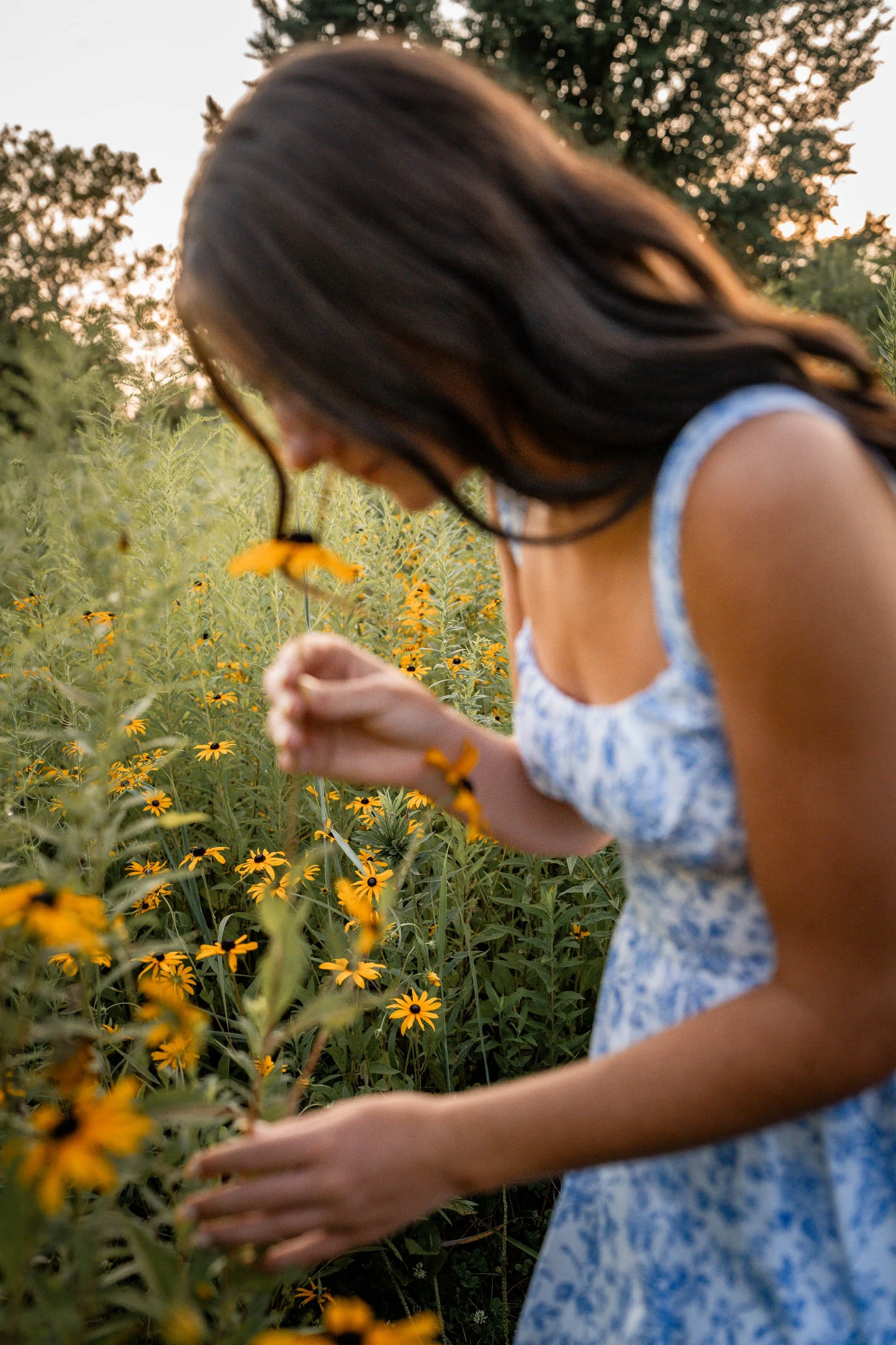 a senior girl wearing a blue and white dress bends over to smell a yellow daisy as her hair begins to fall