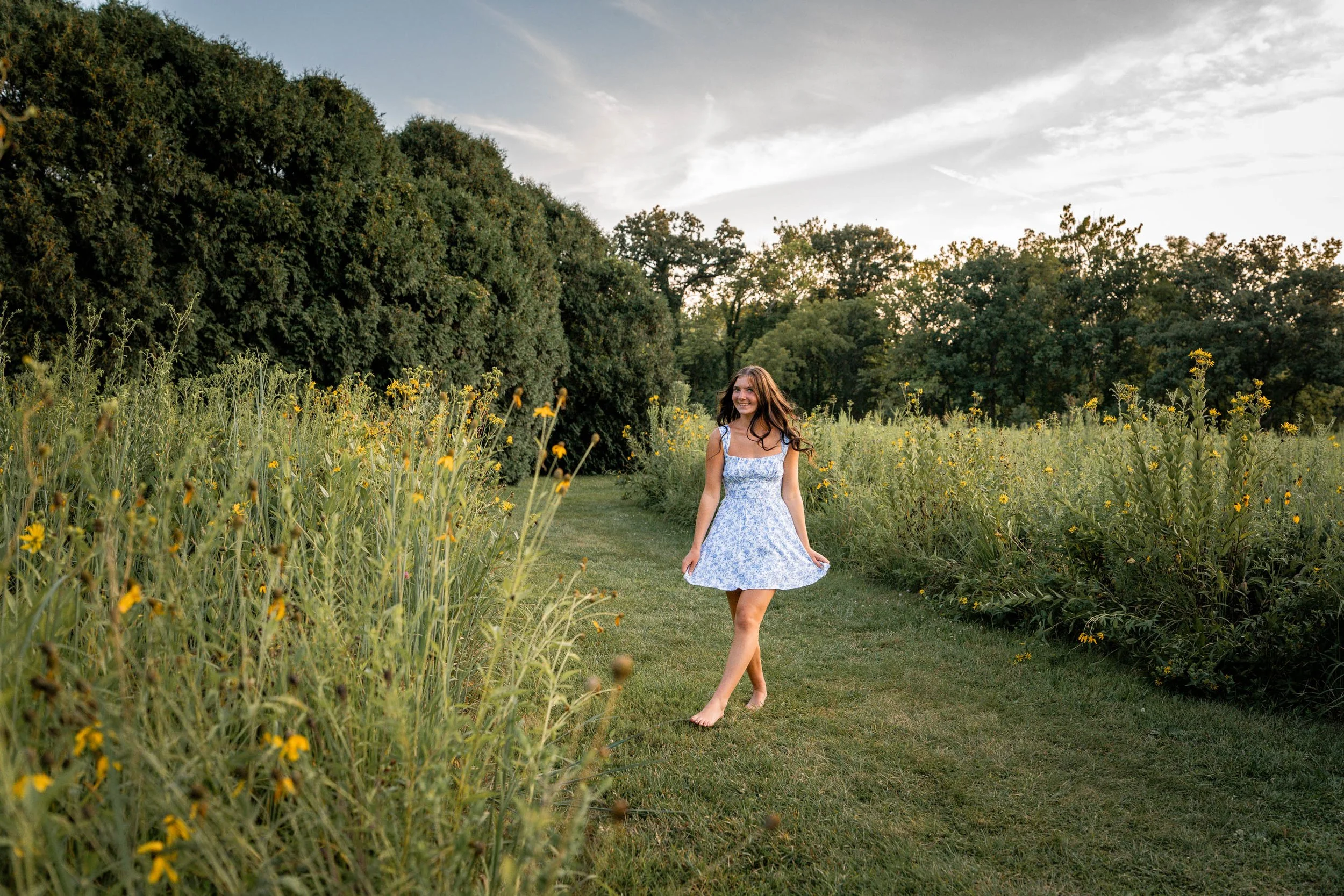 senior girl walks through a field with wildflowers on either side of her