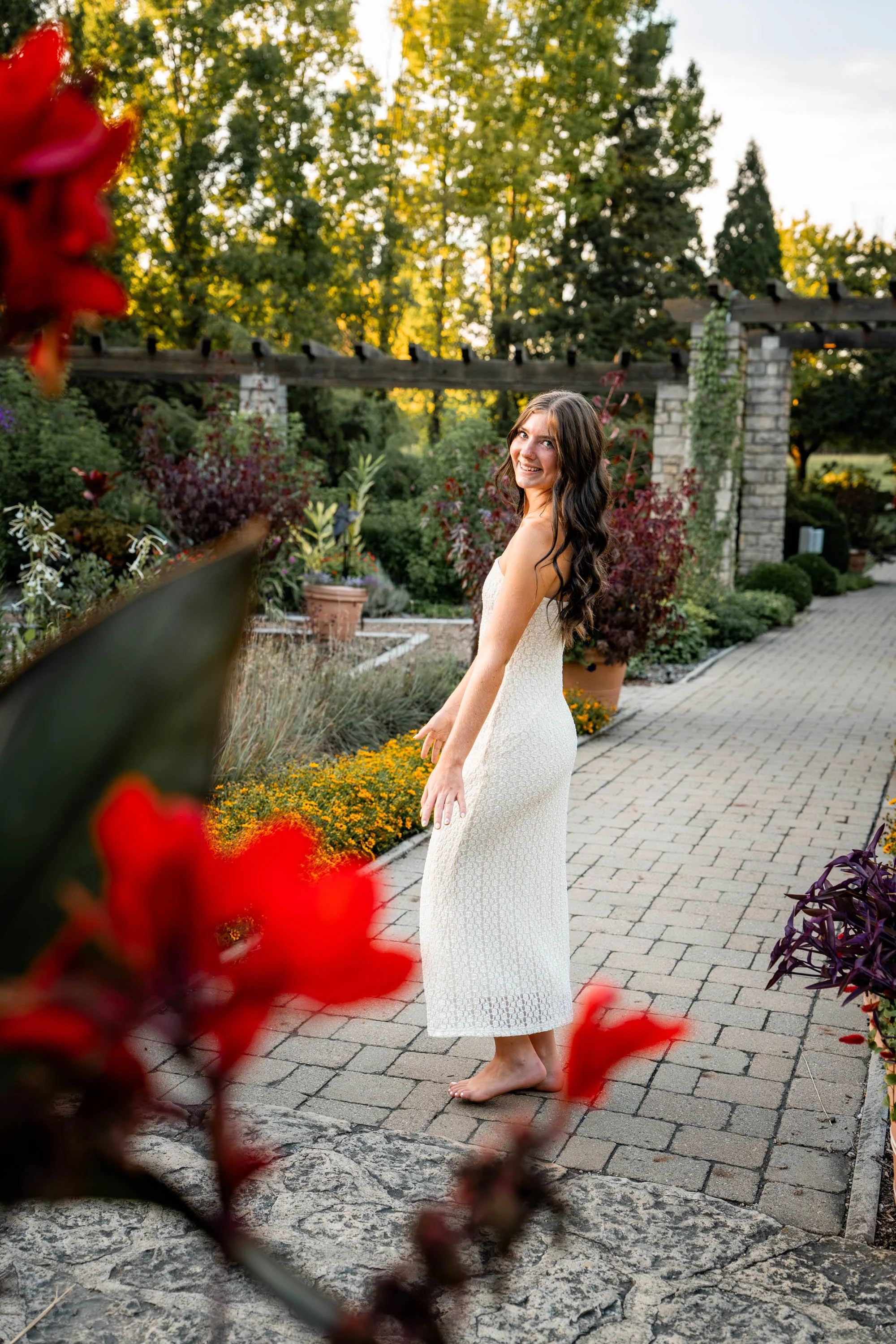 senior girl in a white dress spins as she looks back at the camera through red blooms at Wegerzyn Gardens