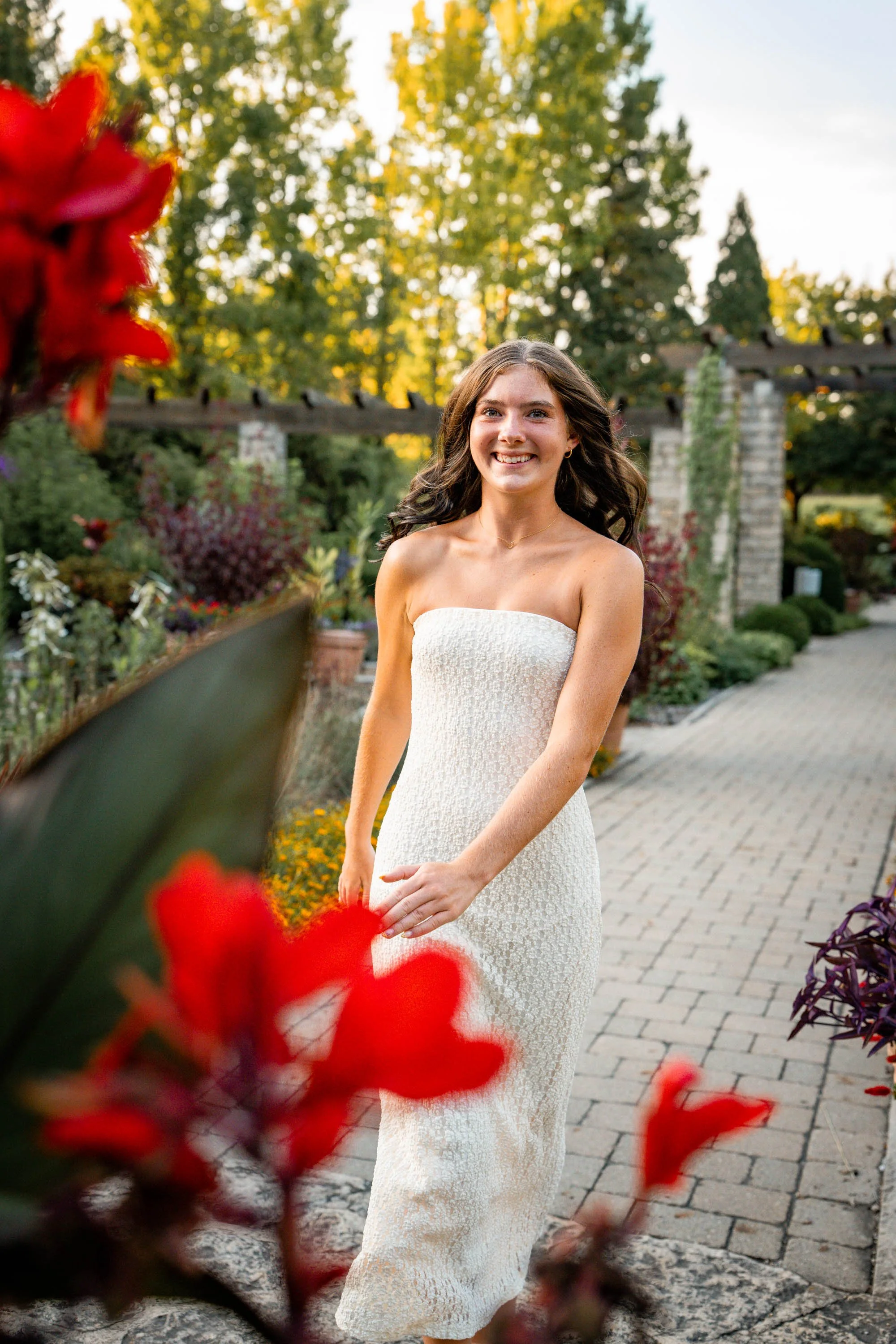 Senior girl skips toward the camera among red blooms with green trees behind her 