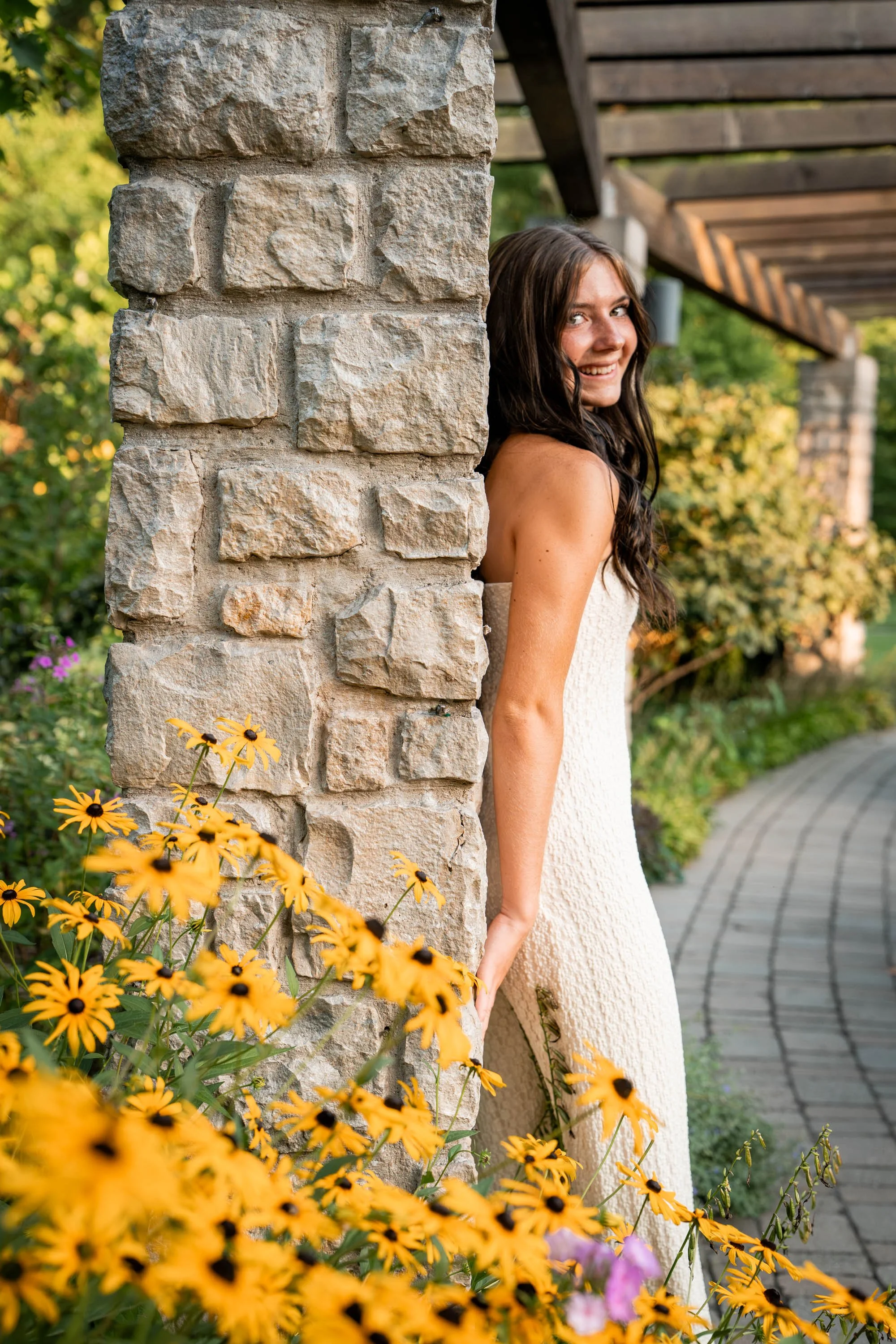 Girl in a white dress leans against a stone pillar behind yellow flowers