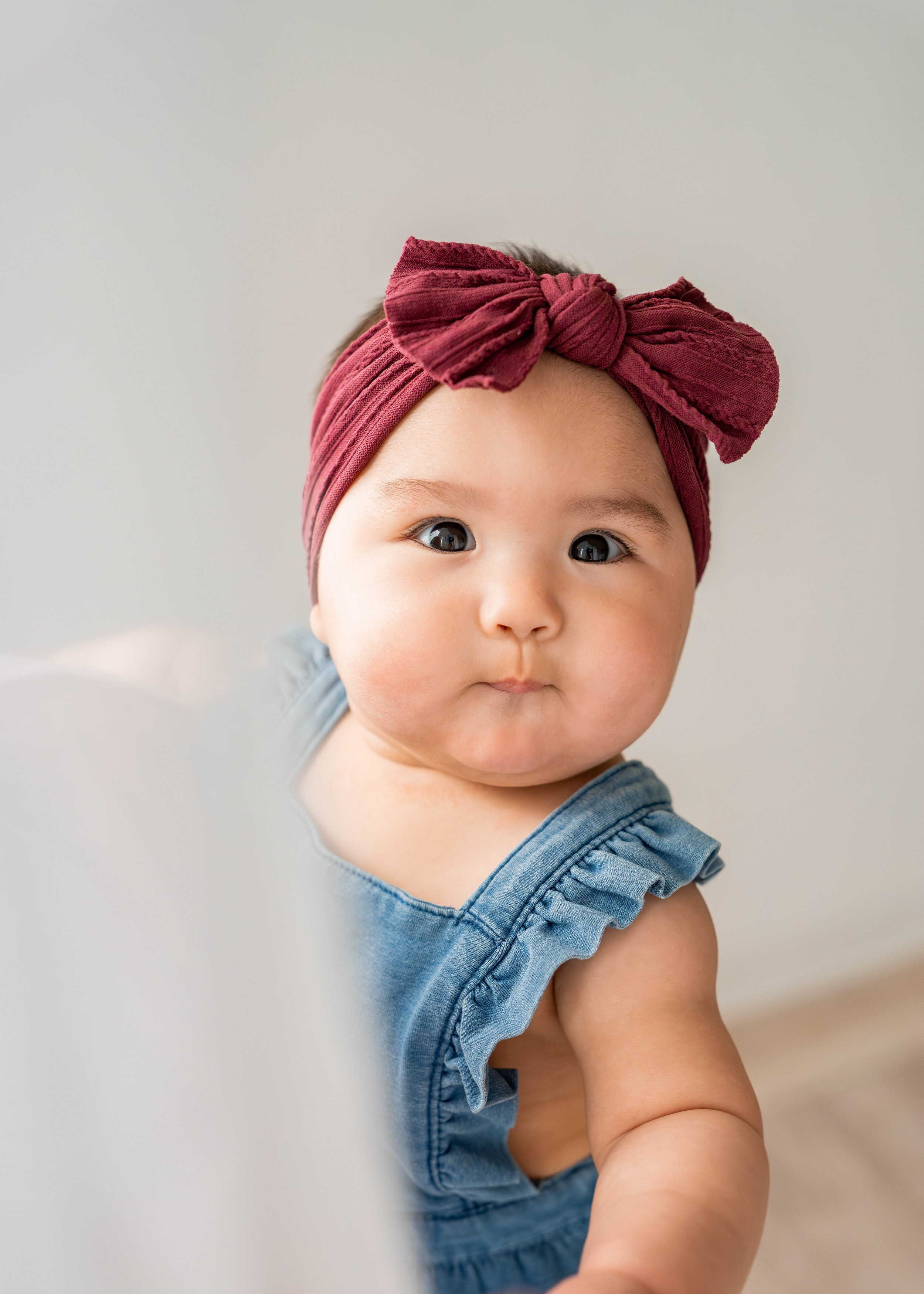 A baby with a blue ruffled dress and a maroon headband with a bow looks curiously at the camera.
