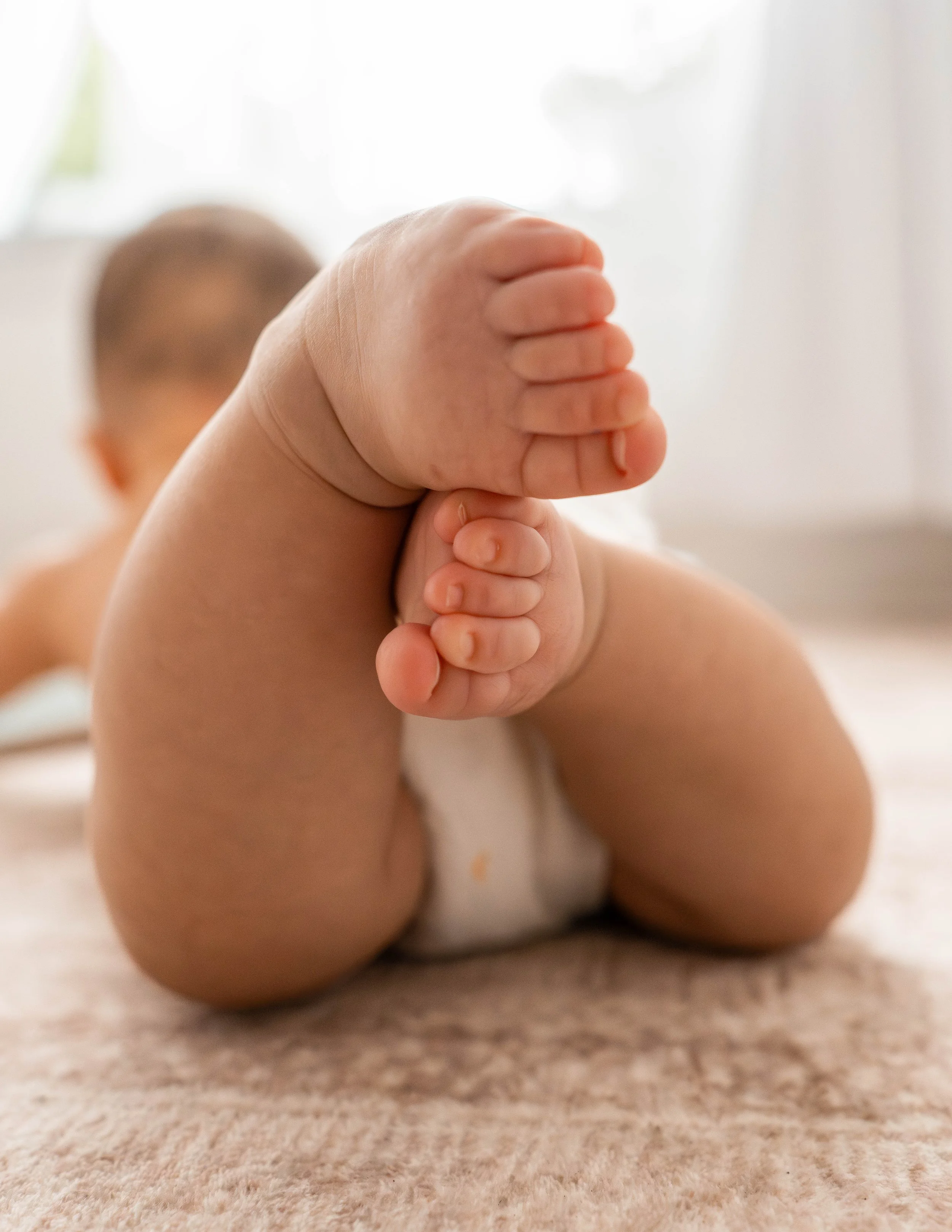 Close-up of a baby's feet and legs on a soft carpet, with the baby lying on their stomach and holding their feet.