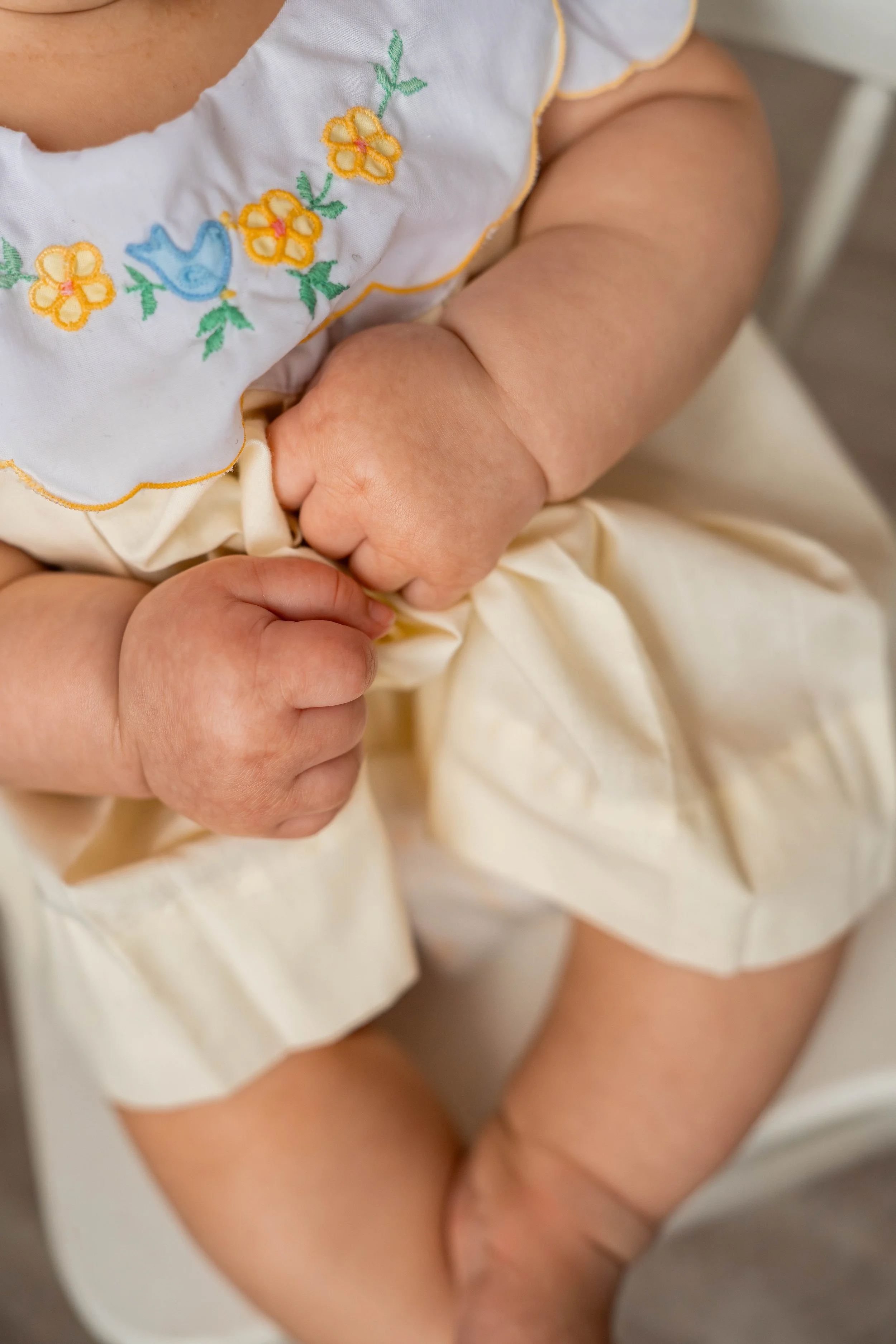 Close-up of a baby's hands holding a cream-colored fabric, wearing a white embroidered dress with yellow and blue details.