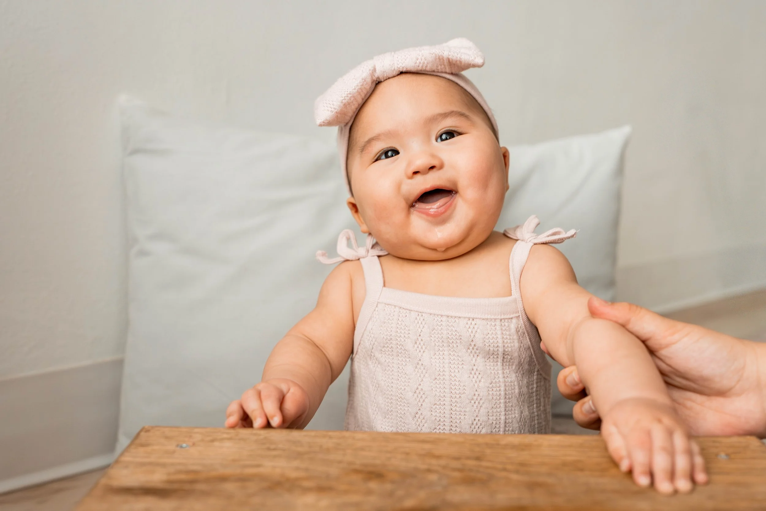 A smiling baby girl with a pink headband and sleeveless dress, sitting at a wooden table, being gently held by an adult's hand.