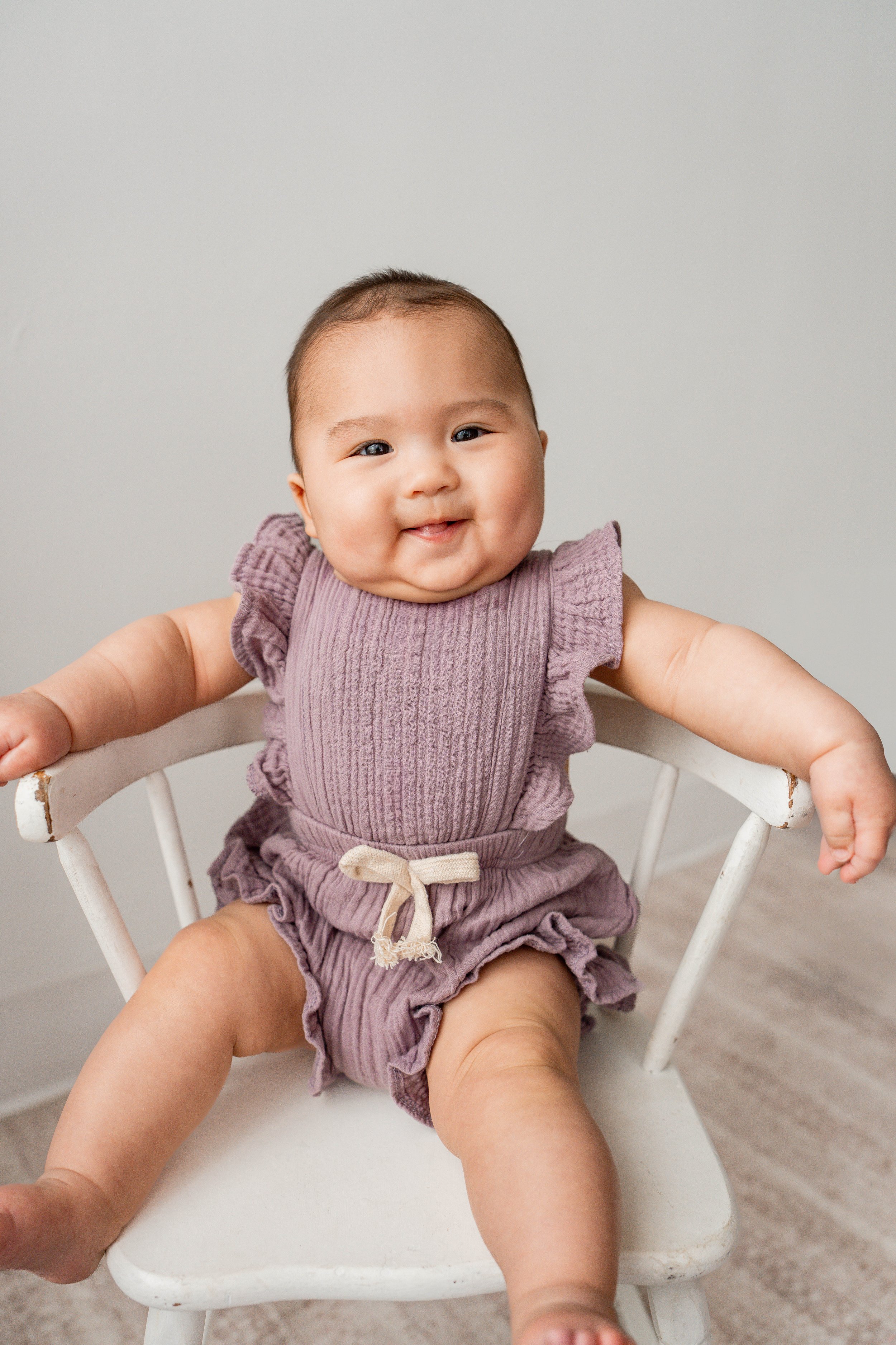 A smiling baby sitting in a white wooden chair with a gray background.