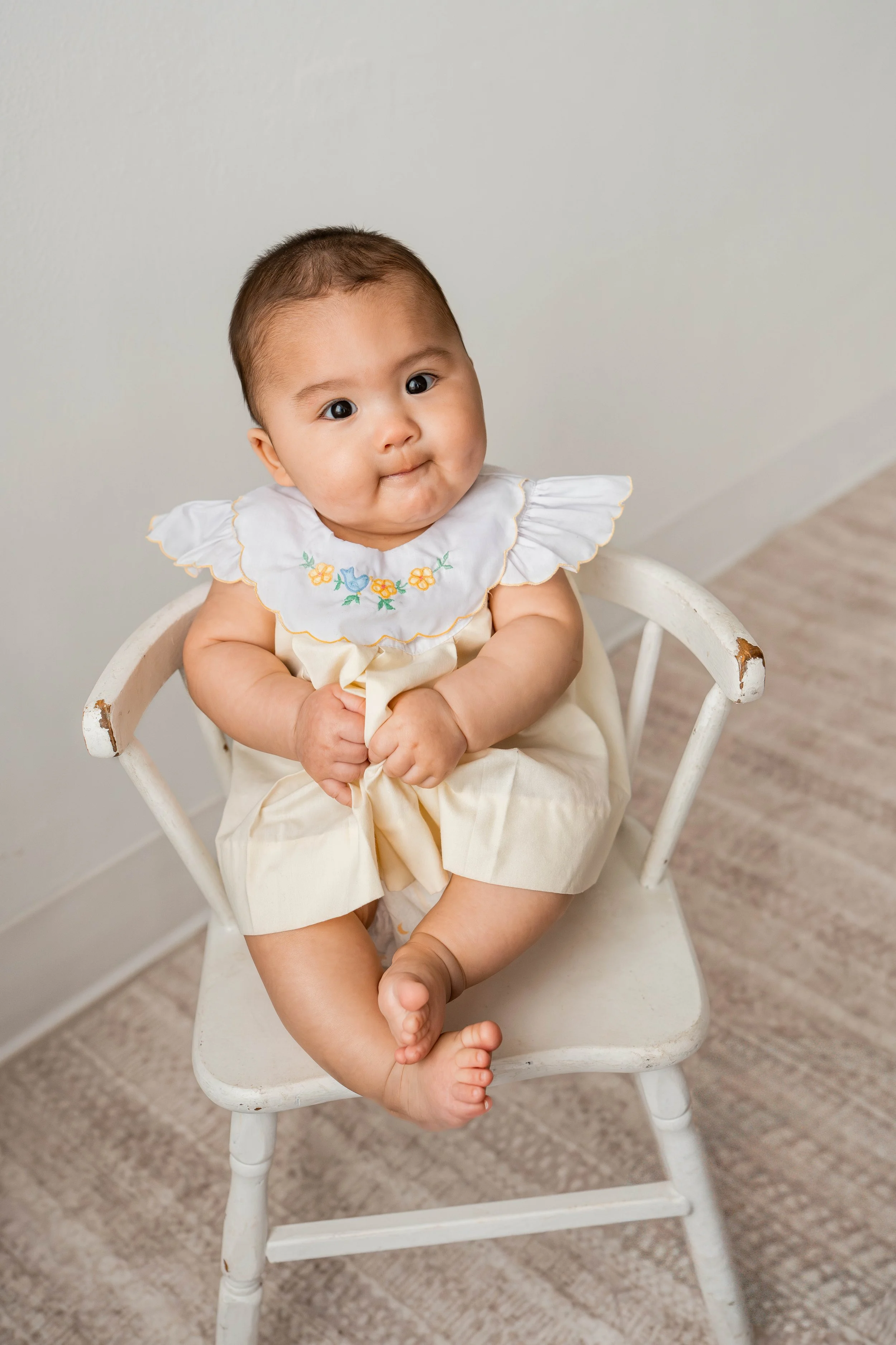 A baby sitting in a vintage white wooden chair, holding the edge of the chair with both hands, wearing a cream-colored outfit with a embroidered white collar, against a plain light-colored wall.