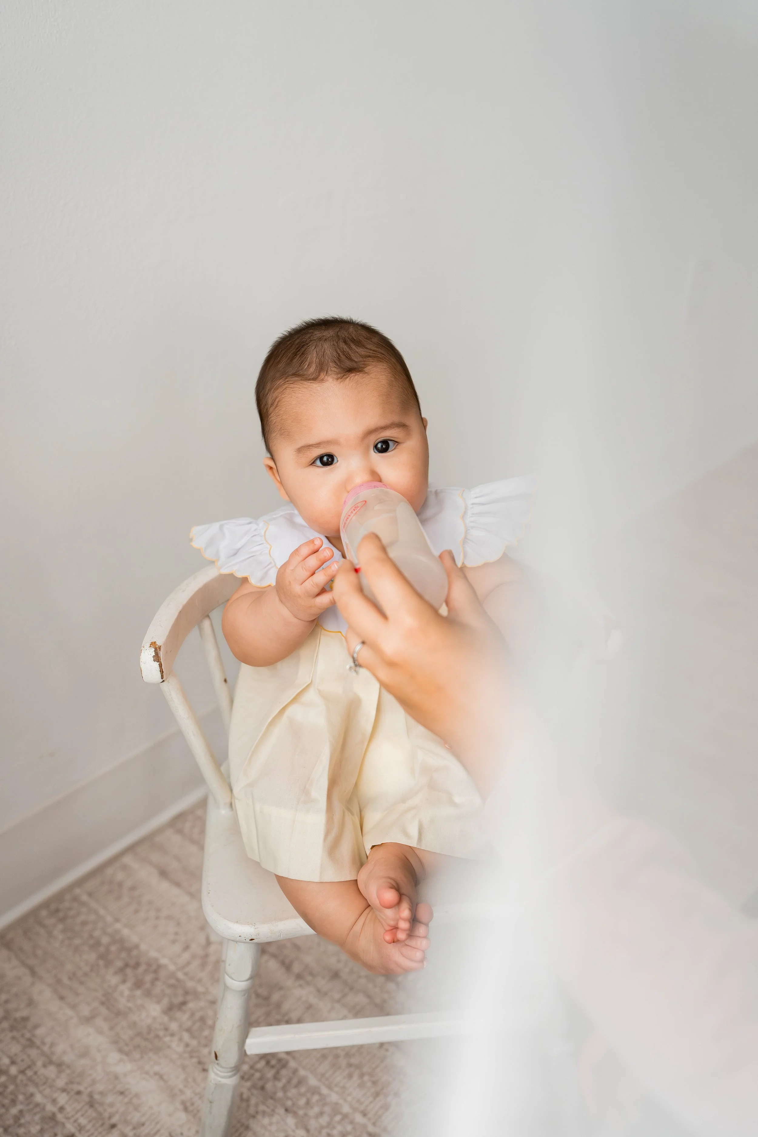 A baby with dark hair and brown eyes sitting on a white wooden chair, drinking from a baby bottle held by an adult hand, against a light-colored background.