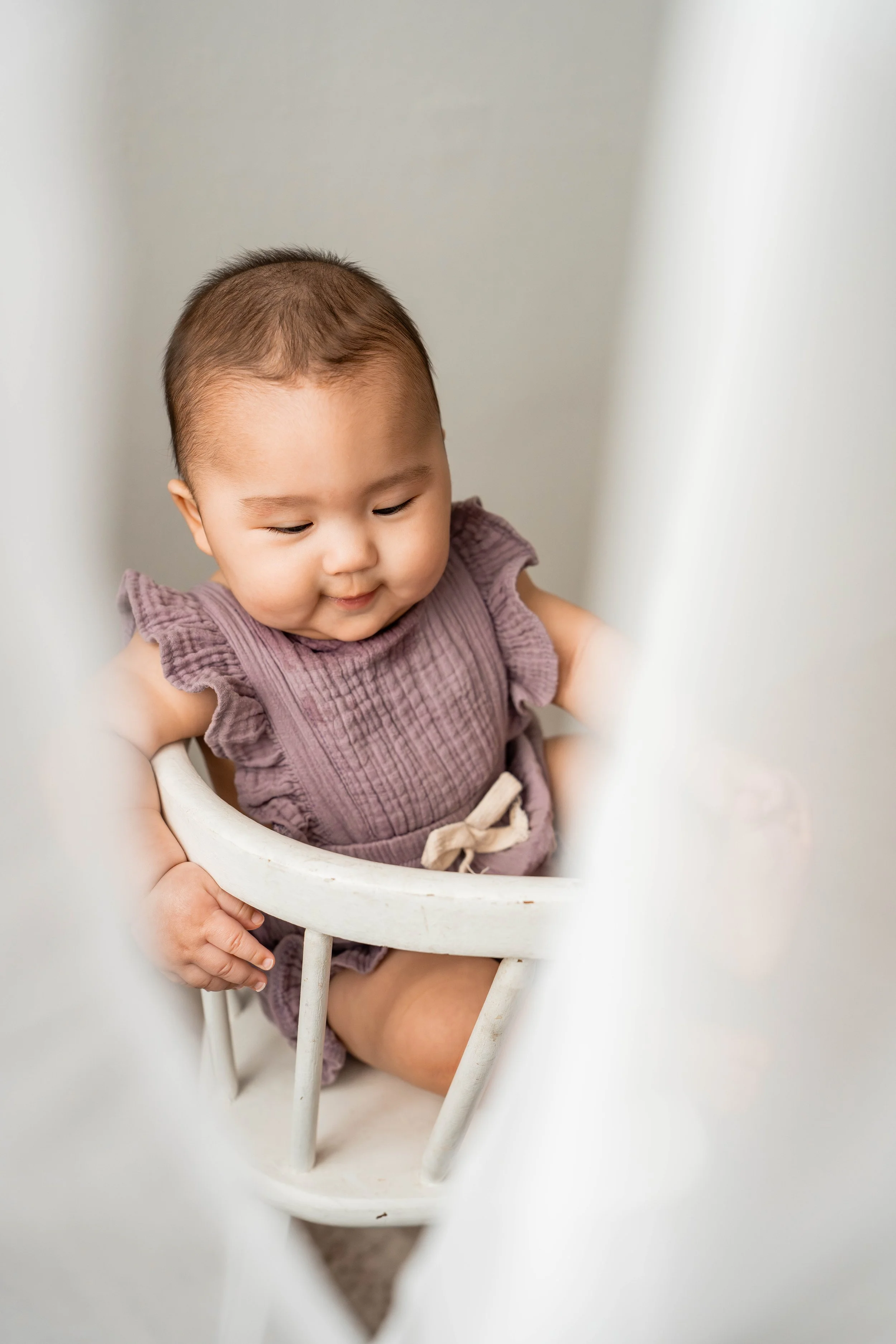 A young Asian girl sitting in a white high chair, wearing a purple dress, looking down and smiling.