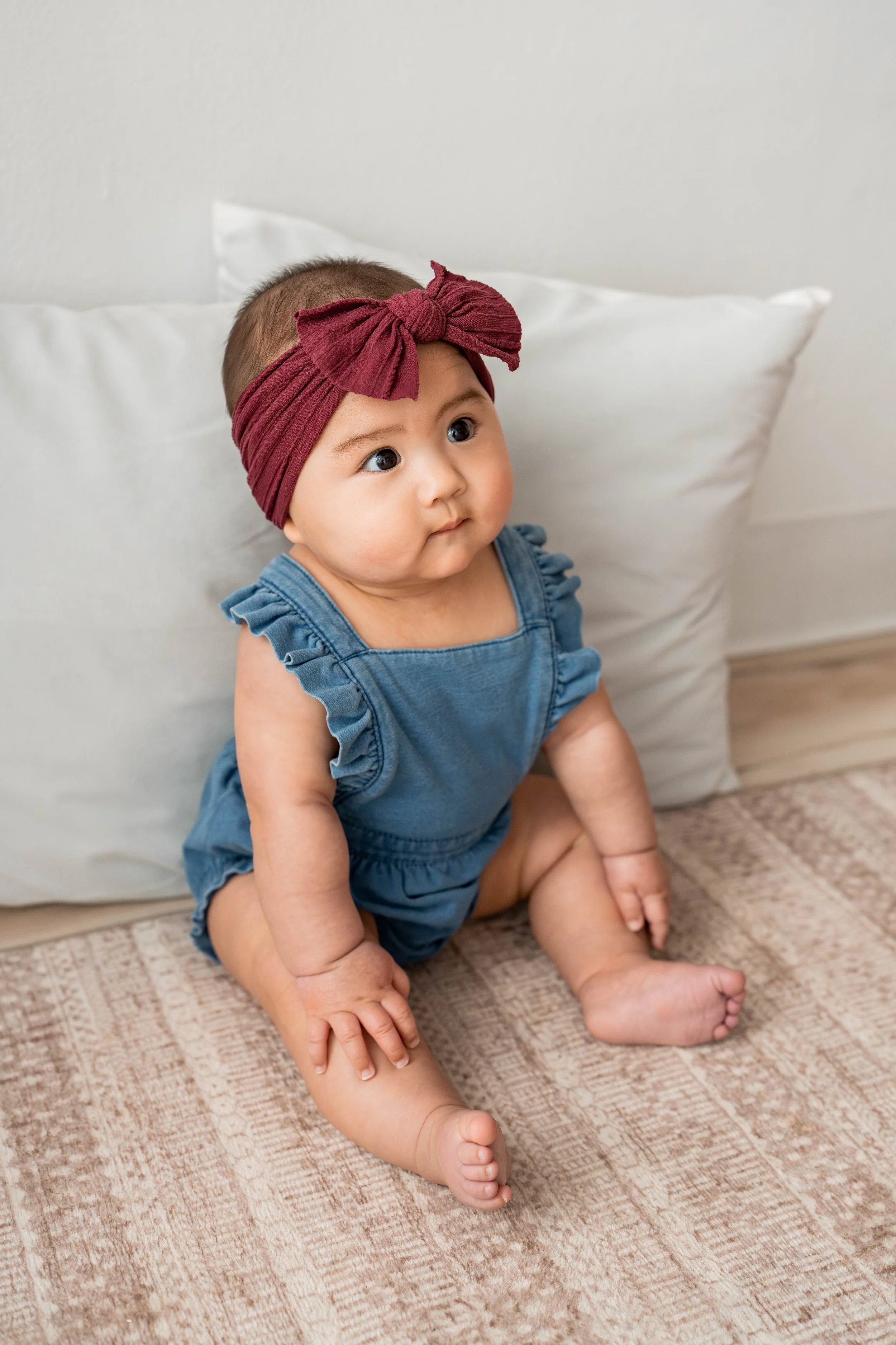 A baby girl with a maroon headband and blue romper sitting on a beige carpet with white pillows behind her in a room with white walls.