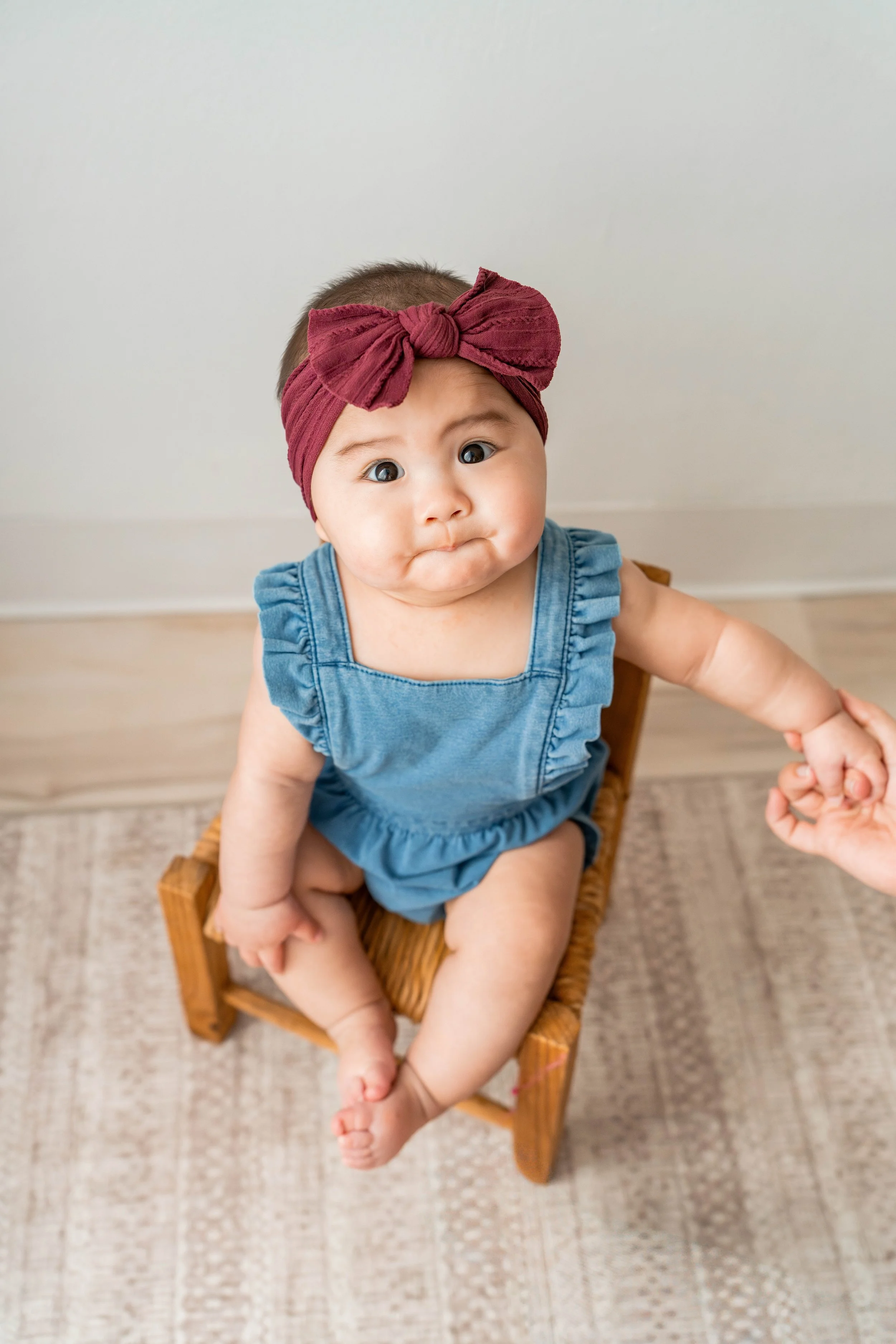 A baby sitting on a small wooden chair, wearing a blue romper and a maroon headband with a bow, holding an adult's hand.