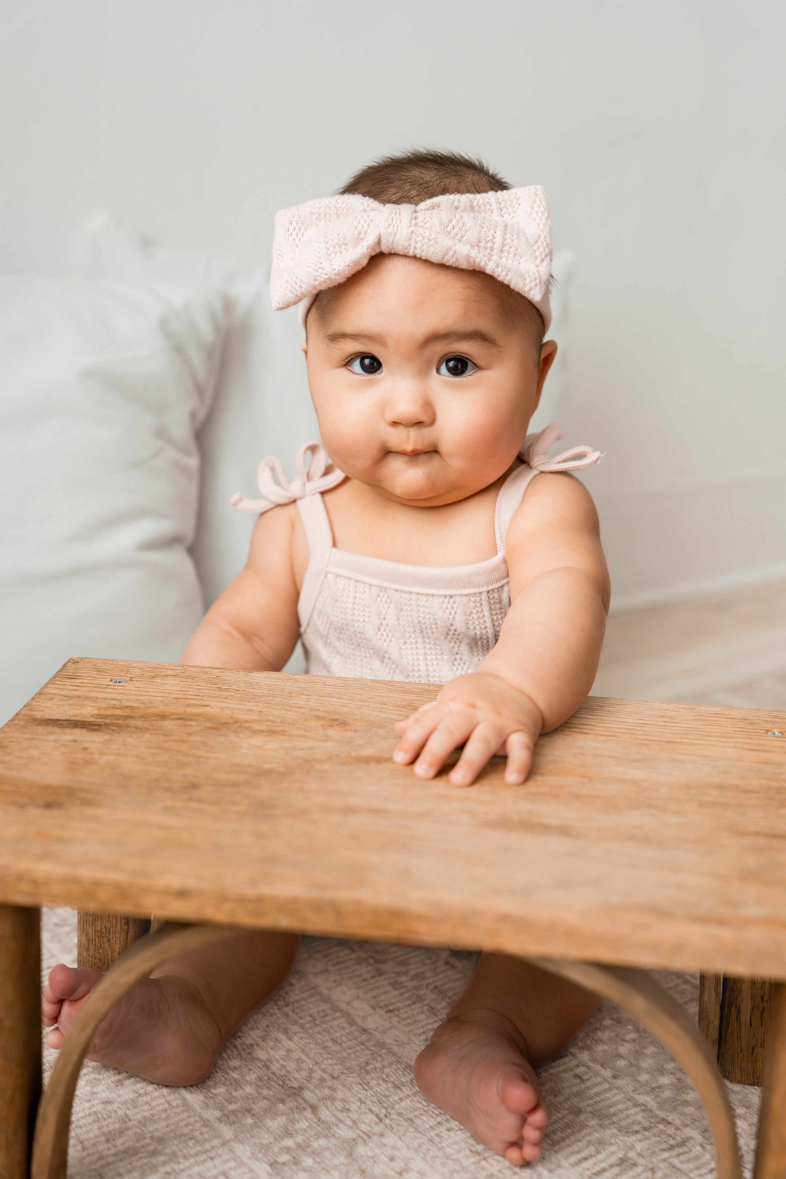 Adorable baby girl with a pink headband and tie-shoulder top, sitting on the floor and reaching over a wooden table, with white pillows in the background.