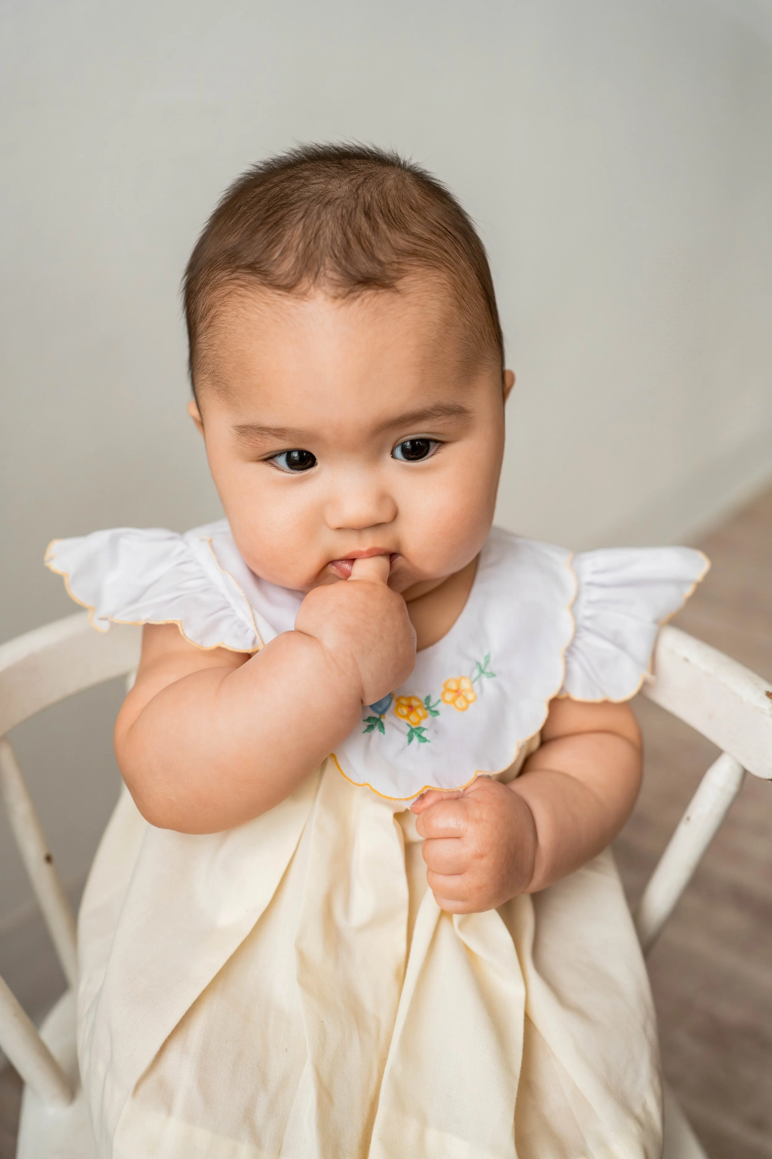A young baby with short hair sitting in a white wooden chair, wearing a white dress with embroidered yellow flowers and yellow trim, with one hand clenched into a fist and the other hand with fingers near mouth.