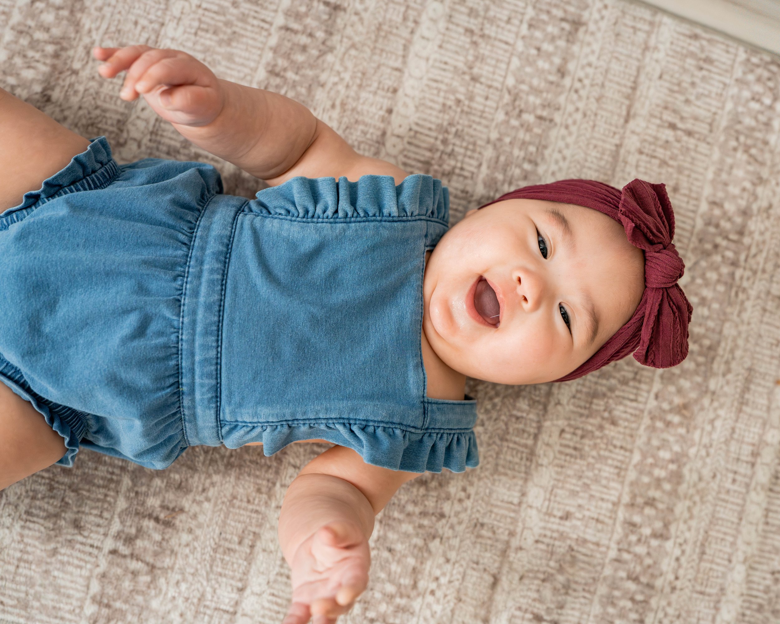 A smiling baby girl lying on a wooden floor, wearing a blue denim romper with ruffled sleeves and a maroon headscarf with a bow.