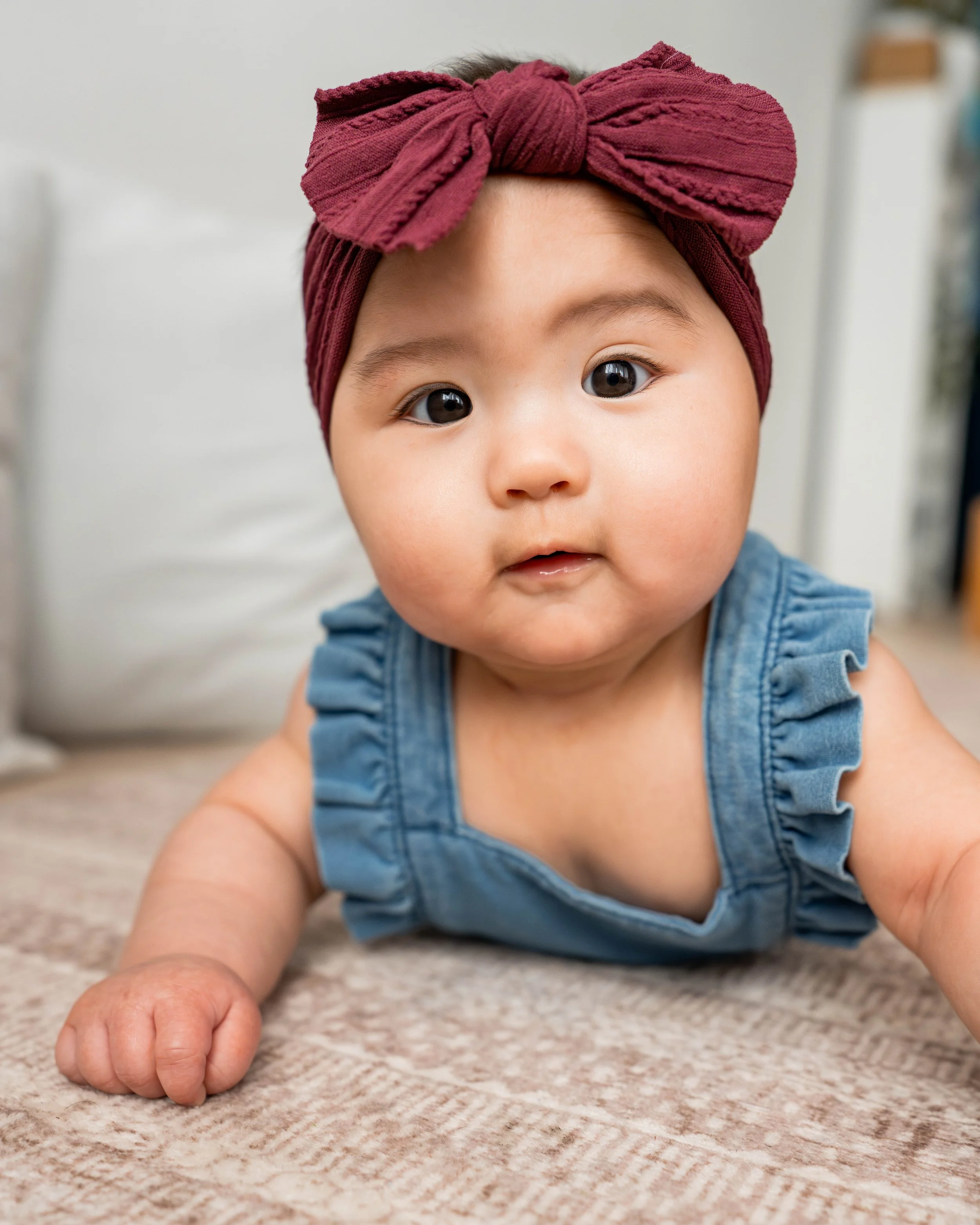 Photo of a cute baby girl with a burgundy headband and a blue ruffled sleeveless top, lying on a beige carpet.