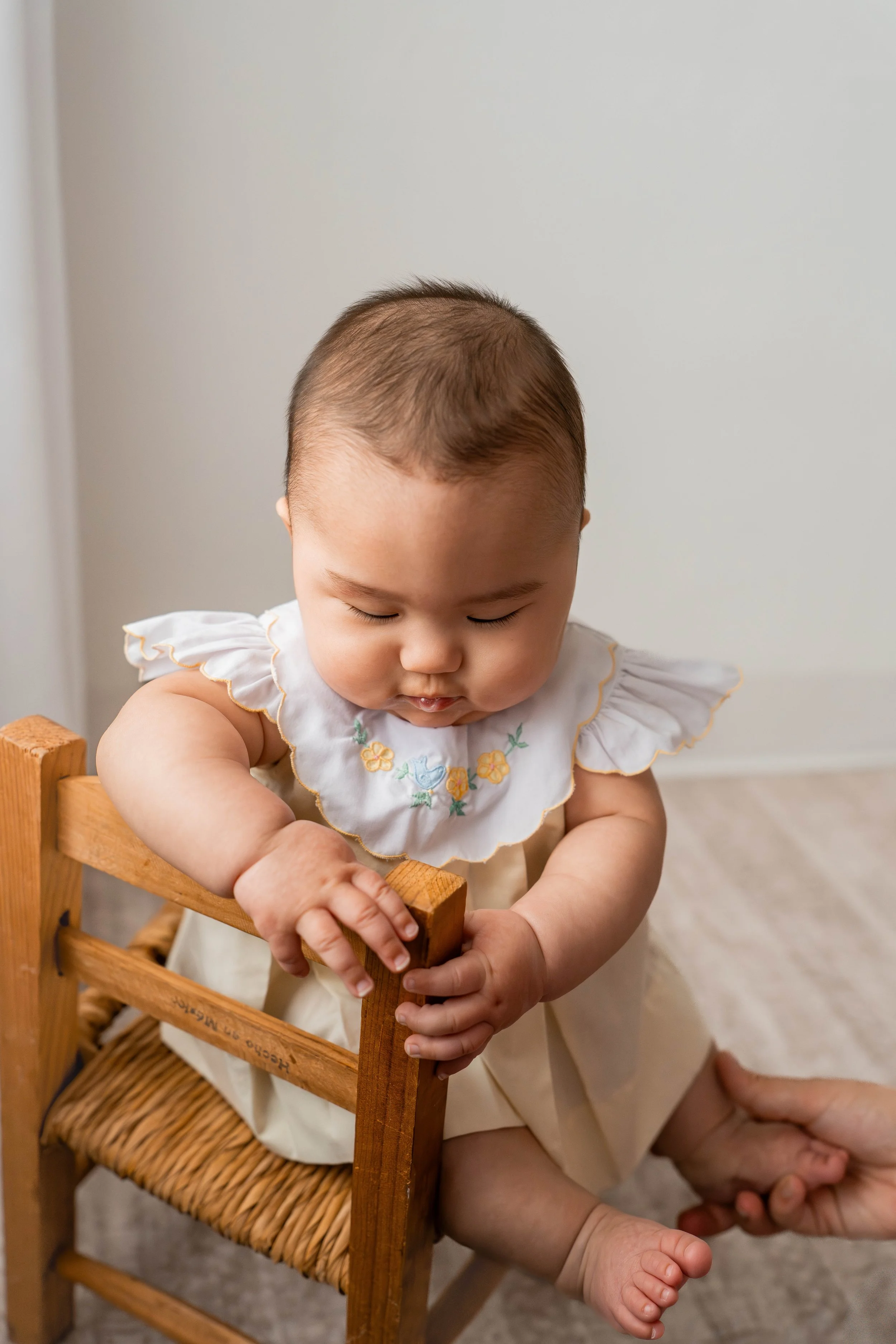 A baby sitting on a small wooden chair, looking down and holding the backrest, with an adult holding one of their feet.