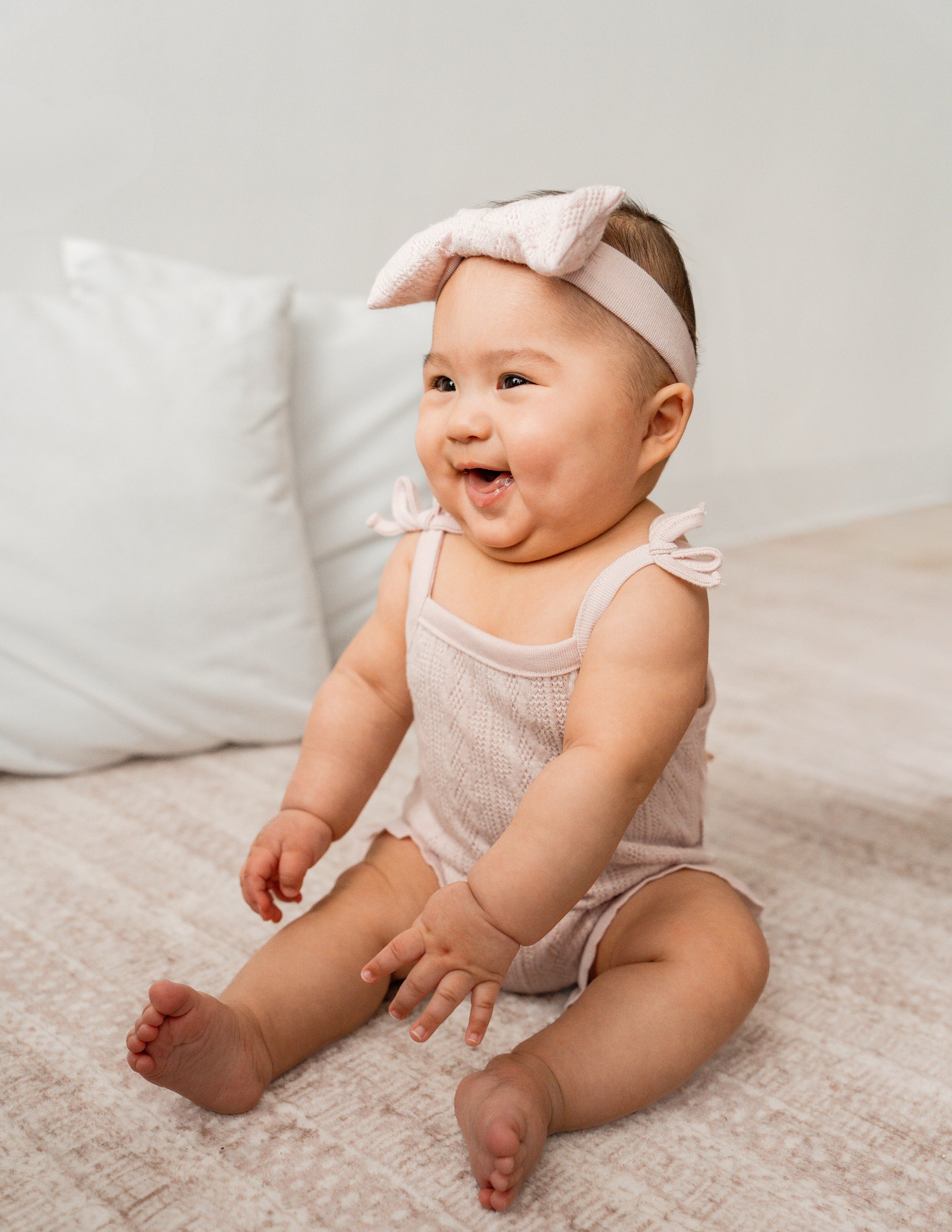 A smiling baby girl sitting on a beige carpet, wearing a light pink romper and a matching headband with a bow, in a bright, simple room.
