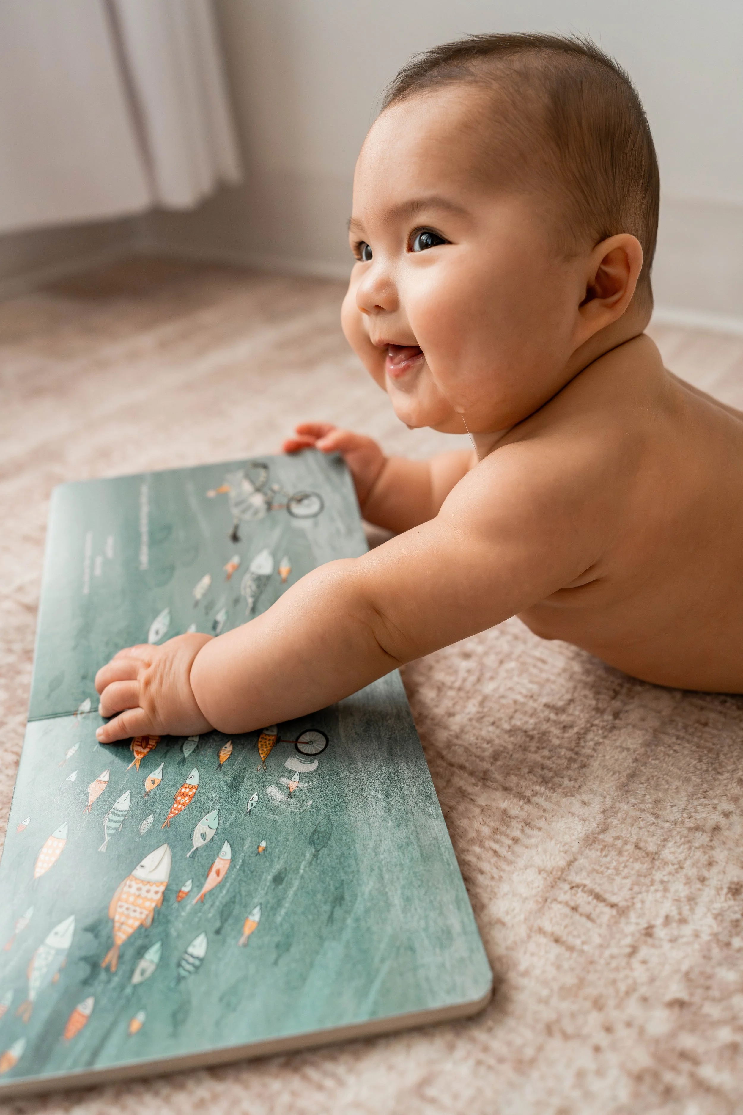 A smiling baby crawling on a carpet and looking to the side while holding a colorful children's book.