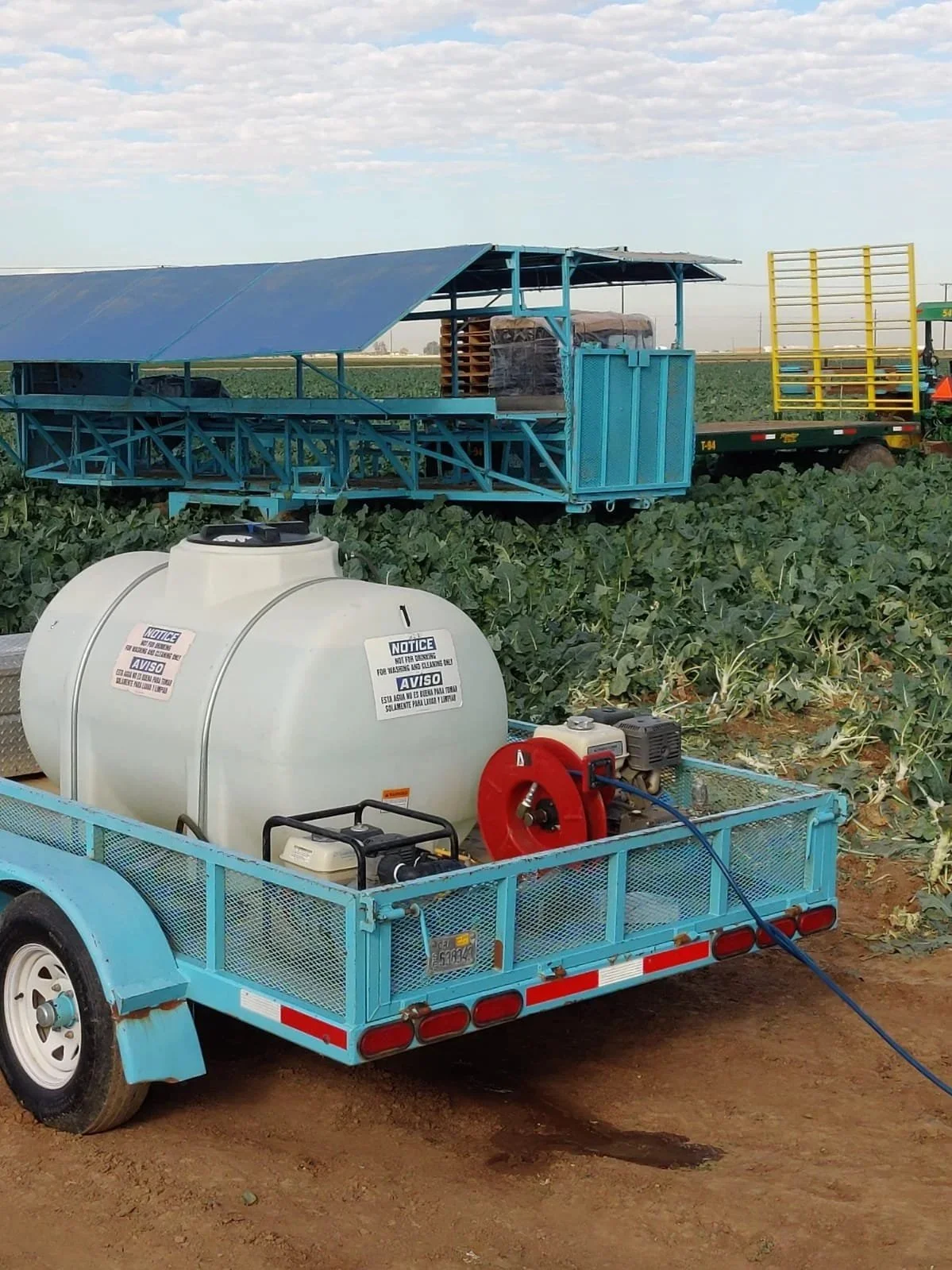 Agricultural spray trailer with tanks and equipment in a green field with blue and yellow trailers in the background.