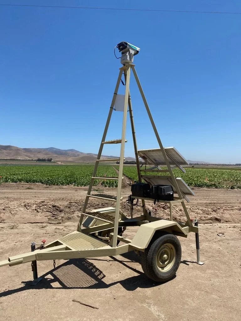Portable solar-powered surveillance camera setup on a trailer in an open field with a mountainous landscape in the background.
