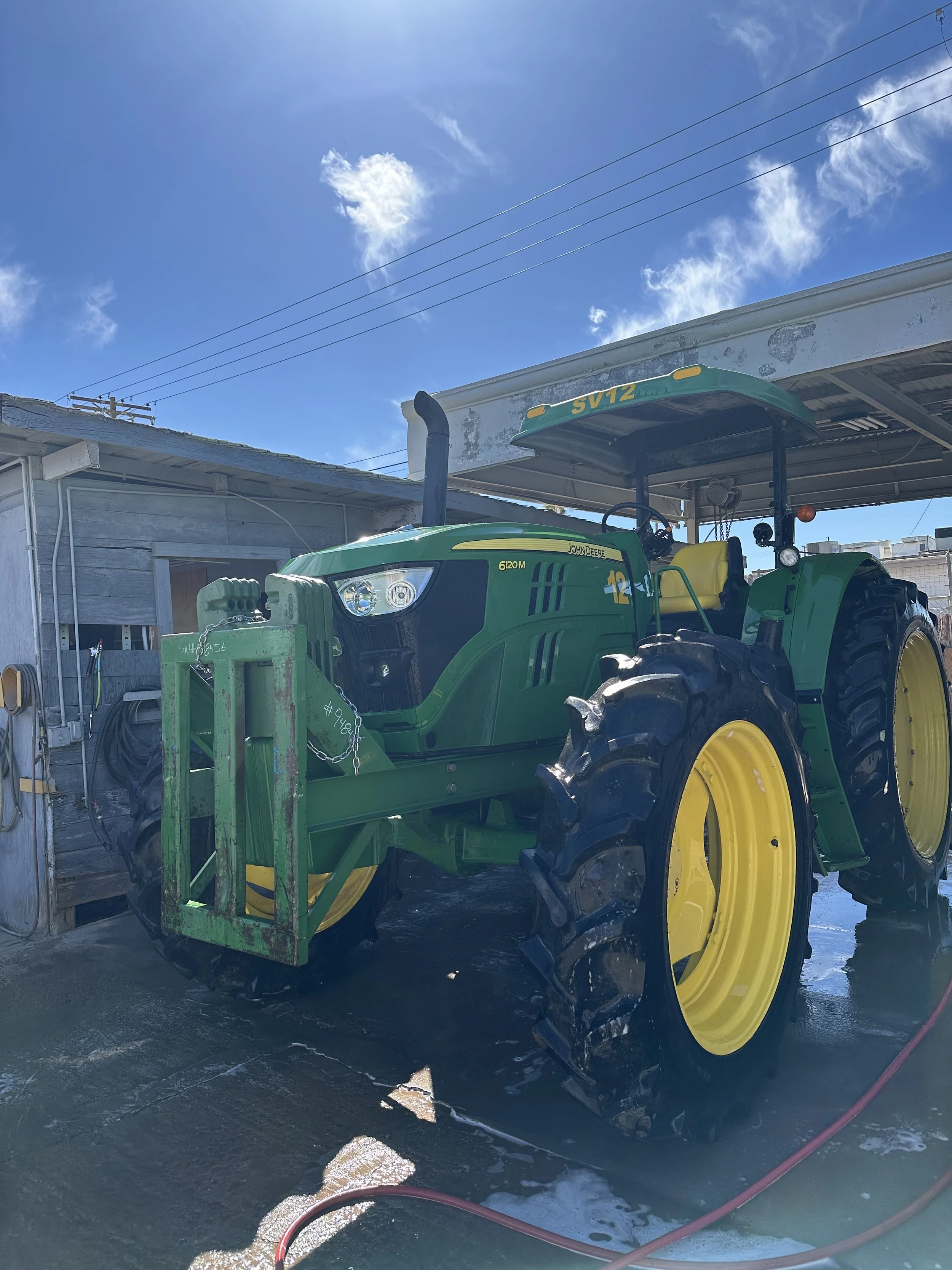 Green John Deere tractor with yellow wheels parked under a metal roof on a concrete surface with soap and water, a blue sky with clouds overhead.