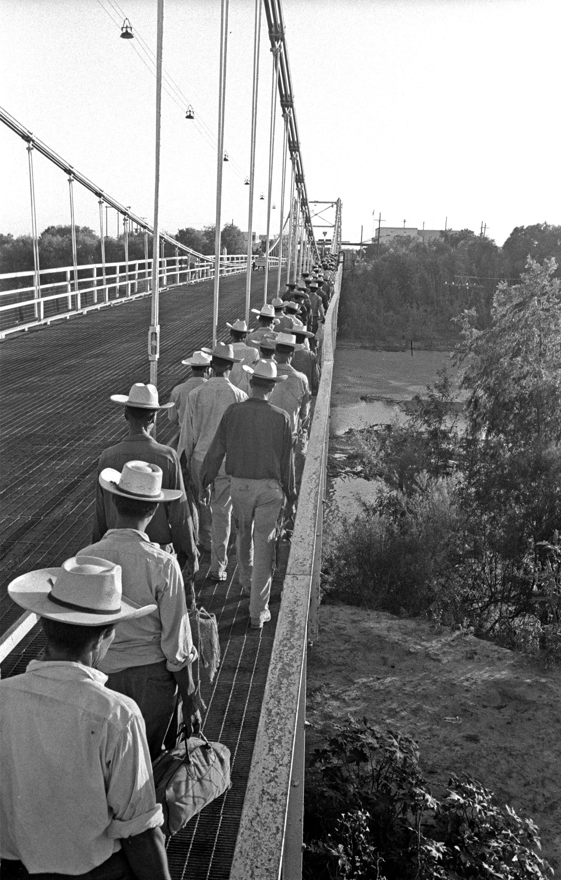 Group of men wearing hats walking in single file across a suspension bridge.