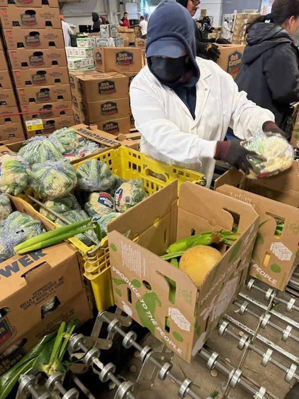 A worker in a grocery store wearing a white coat, face mask, cap, and gloves, packing vegetables into a box. There are boxes of produce, including broccoli, leeks, and a melon, in front of her.
